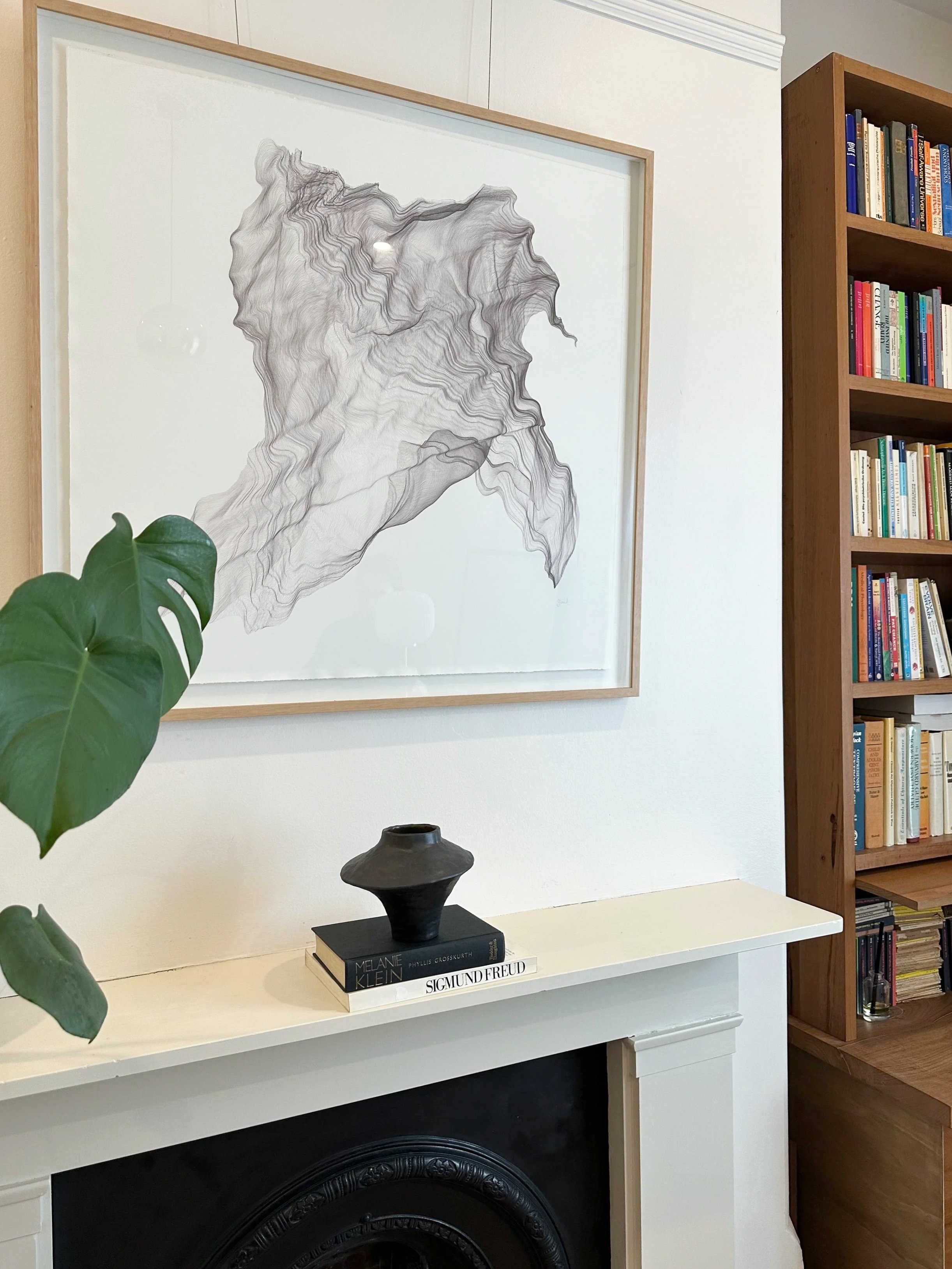 A living room corner with a white fireplace mantel, a black and white abstract artwork on the wall, a wooden bookshelf filled with books, and a white table with a black sculpture and books on top. A green leafy plant partially visible on the left.