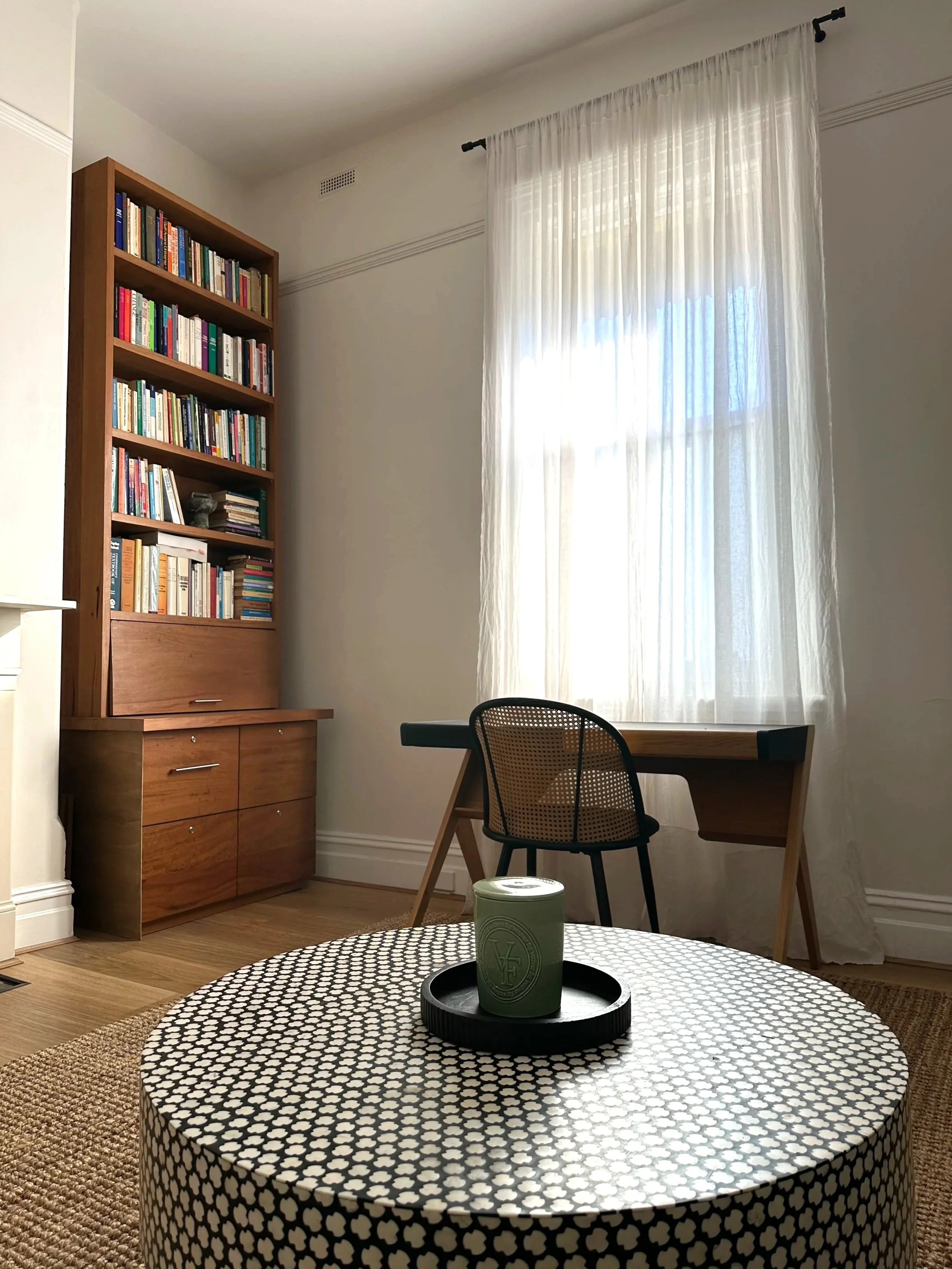 A cozy room with a round table covered in a black and white clover pattern tablecloth, a green candle on a black tray, a wooden desk at the window with a black mesh-backed chair, a wooden bookshelf filled with books, and sheer white curtains allowing