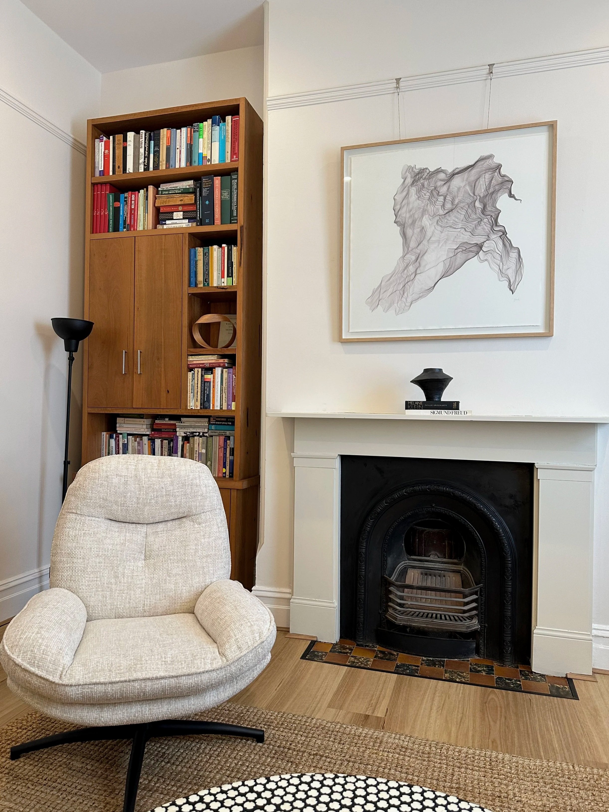 A cozy psychotherapy consulting room with a beige armchair on a brown rug, a tall black floor lamp, a wooden bookshelf filled with books, a white fireplace with a black interior, and a framed abstract line drawing on the wall.