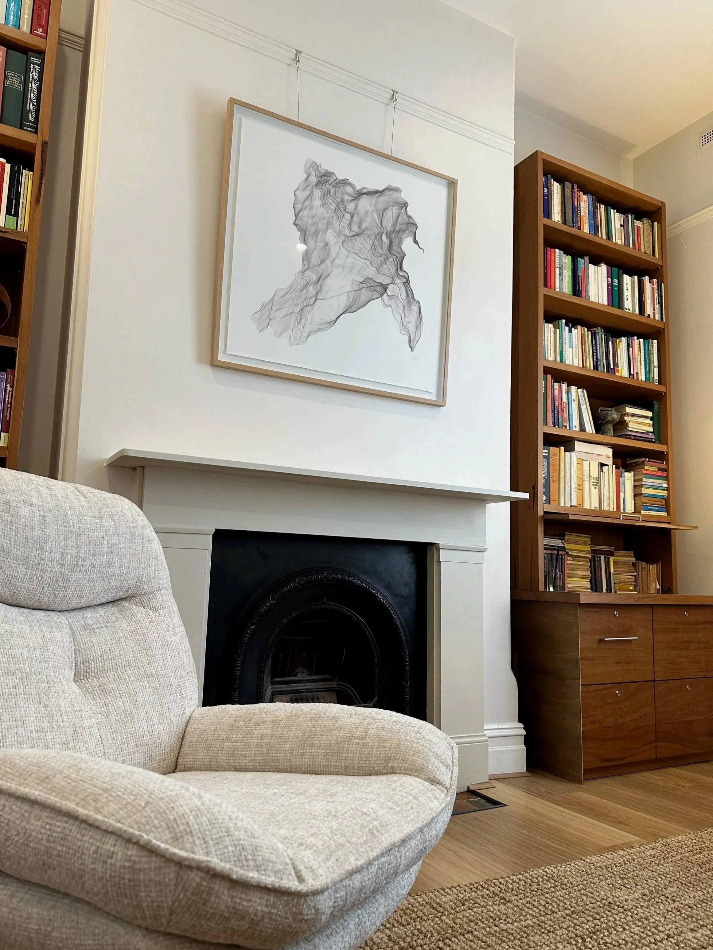 A cozy living room corner featuring a beige sofa with plush cushions, a white fireplace with a decorative black insert, a large abstract map artwork hanging above the fireplace, and two wooden bookshelves filled with colorful books.
