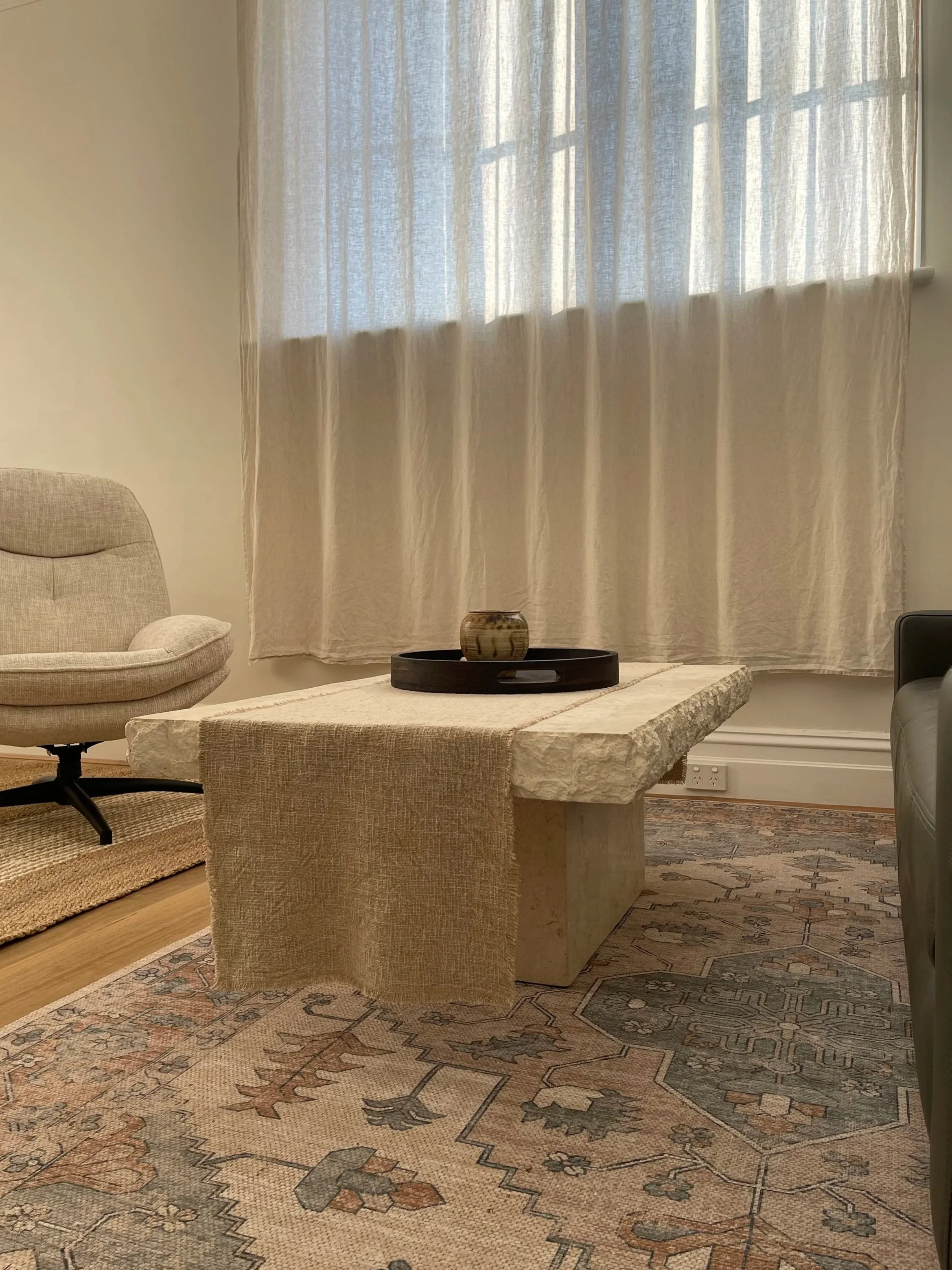 A beige upholstered armchair with a curved back and armrests, a large stone coffee table with a small decorative vase, a patterned rug, a window with white semi-transparent curtains, and a black side table with a white electrical outlet visible on th