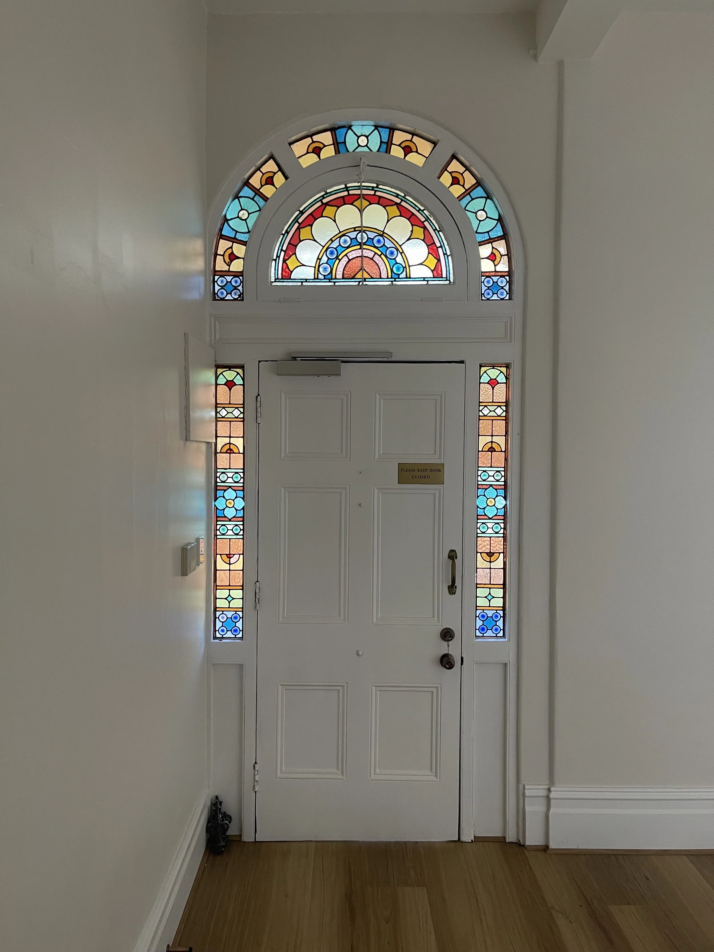White front door with stained glass window above and on sides, with a staircase visible on the right. There is a sign on the door that reads 'Please keep door closed' and a shoe rack with shoes on the floor to the left.
