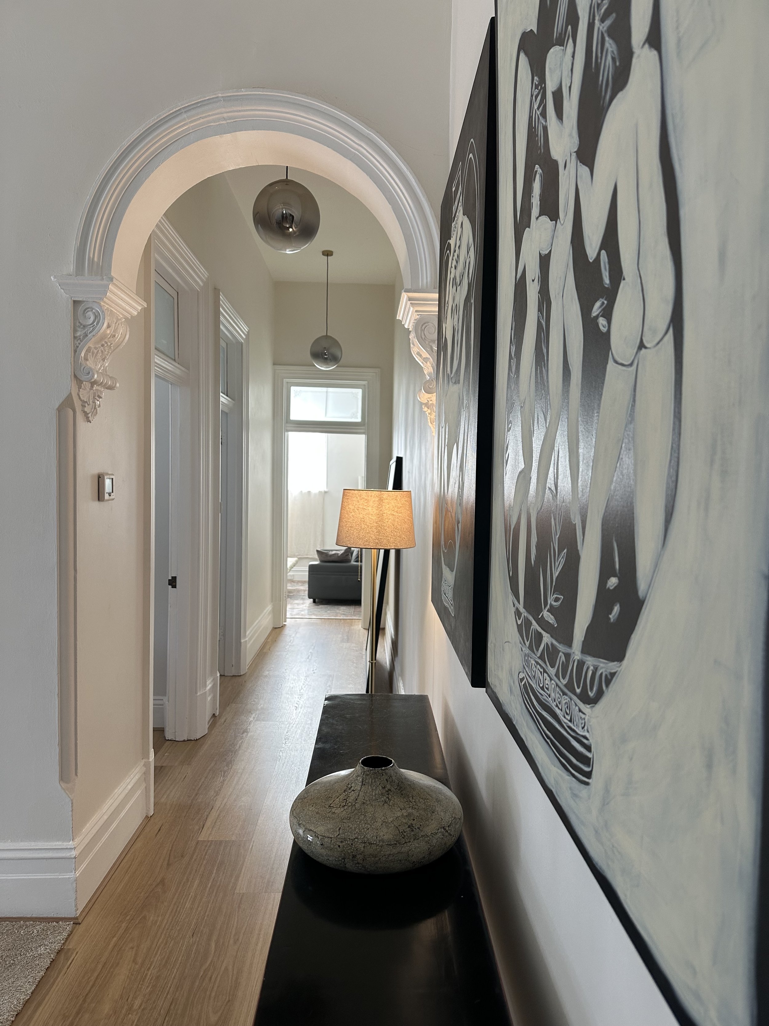 Hallway with hardwood floor, decorative archways, modern hanging pendant lights, a large abstract black-and-white painting on the right wall, a table lamp, and a ceramic vase on a black table.
