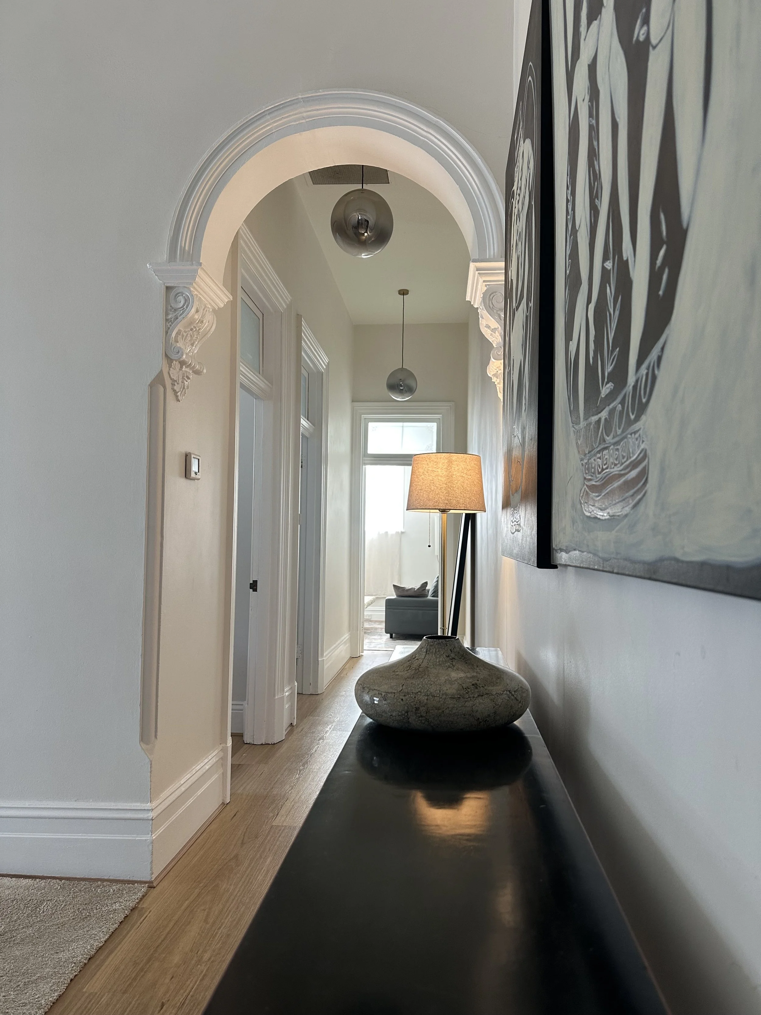 Interior hallway view with decorative wall molding, two spherical pendant lights hanging from the ceiling, a floor lamp with a beige shade, and abstract art on the wall. At the end of the hallway, a sofa with pillows and a large window is visible.
