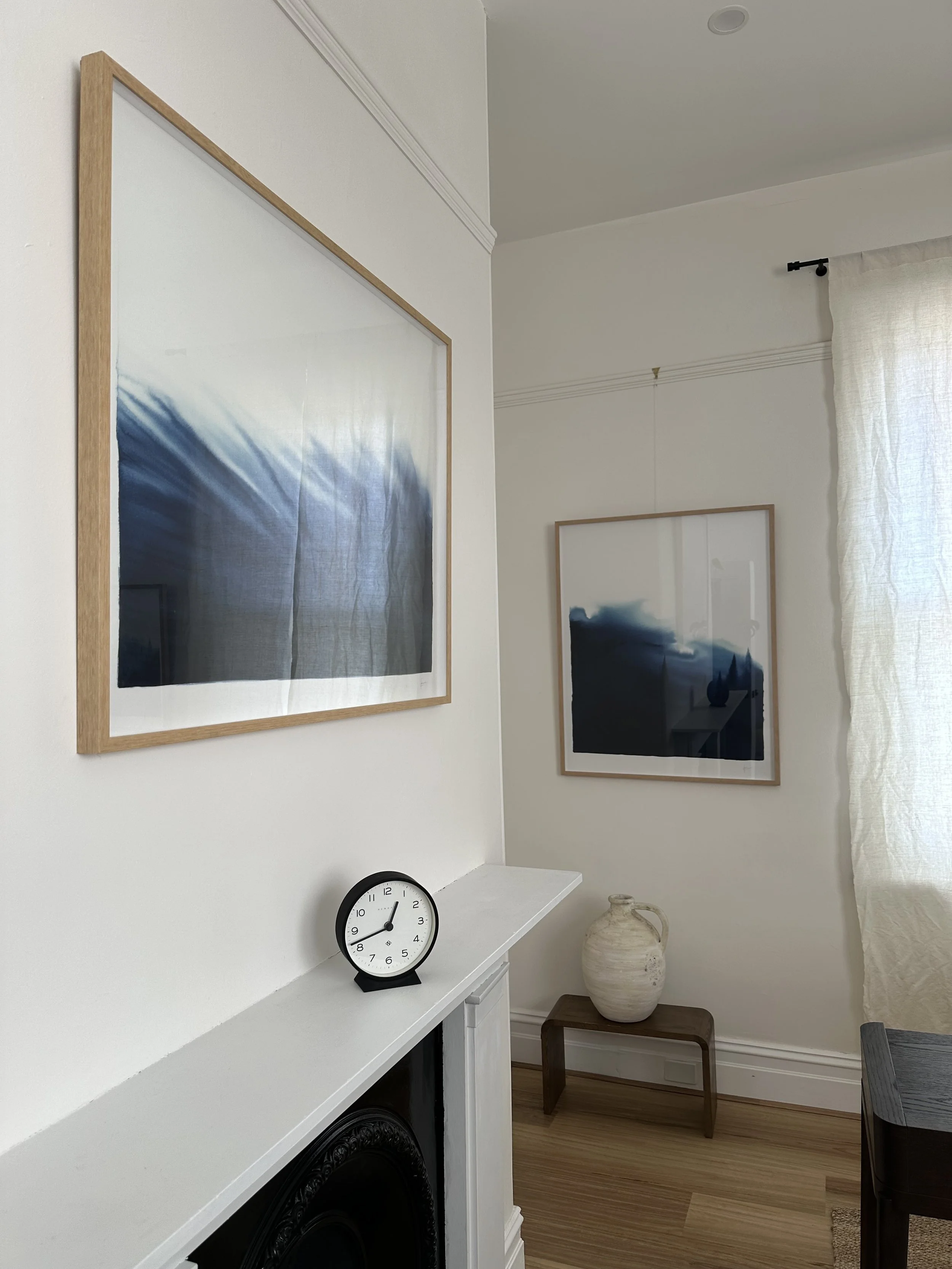 A corner of a psychotherapy consulting room with white walls, decorated with framed artwork, a clock on the mantel, and a large ceramic vase on a small wooden table, with a cream-colored curtain hanging beside a window.