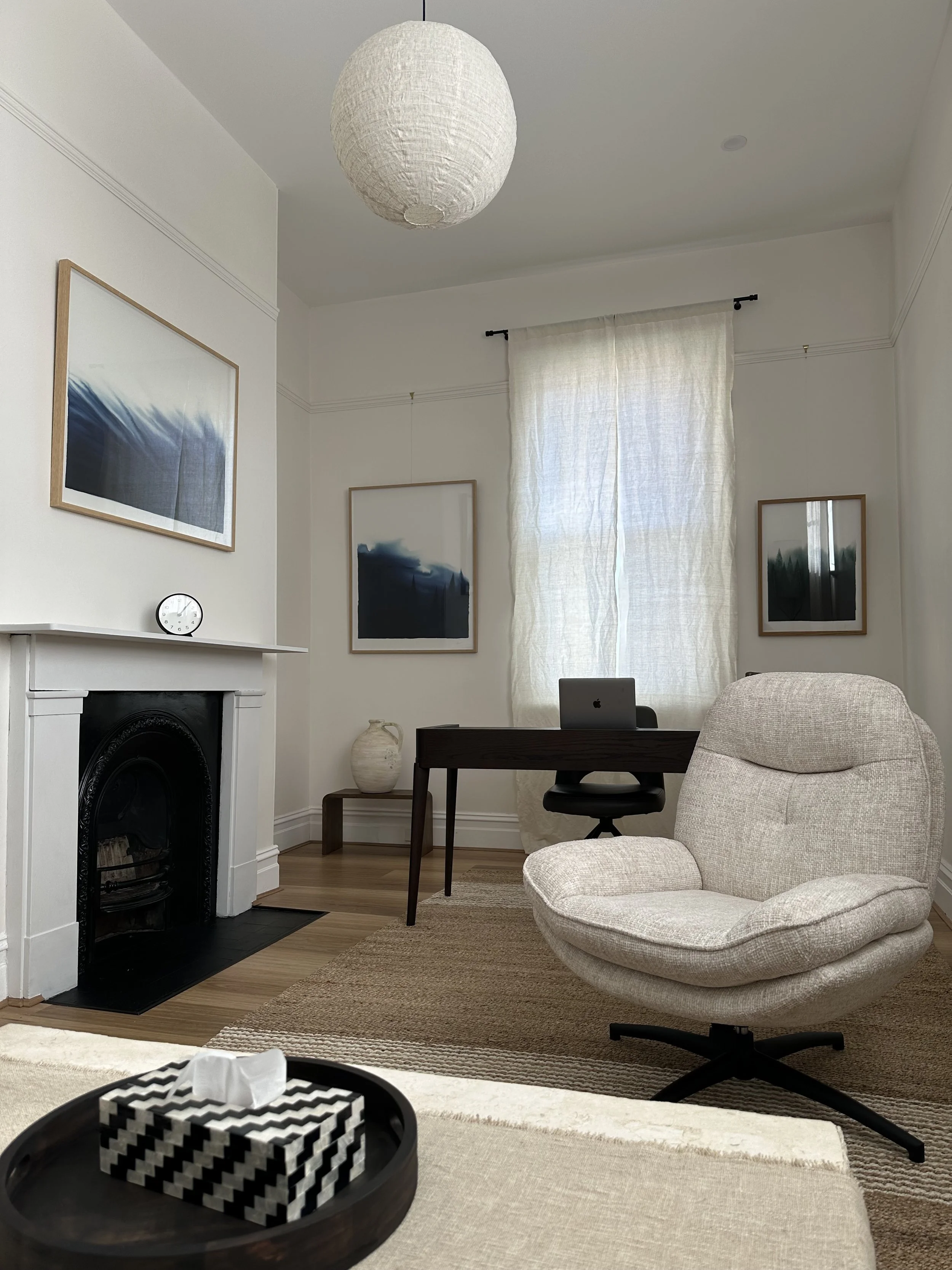 A cozy living room with a white fireplace, framed landscape artwork, a beige armchair, and a small workspace with a desk, laptop, and decorative vase. Light filters through curtains on a window, and a paper lantern hangs from the ceiling.