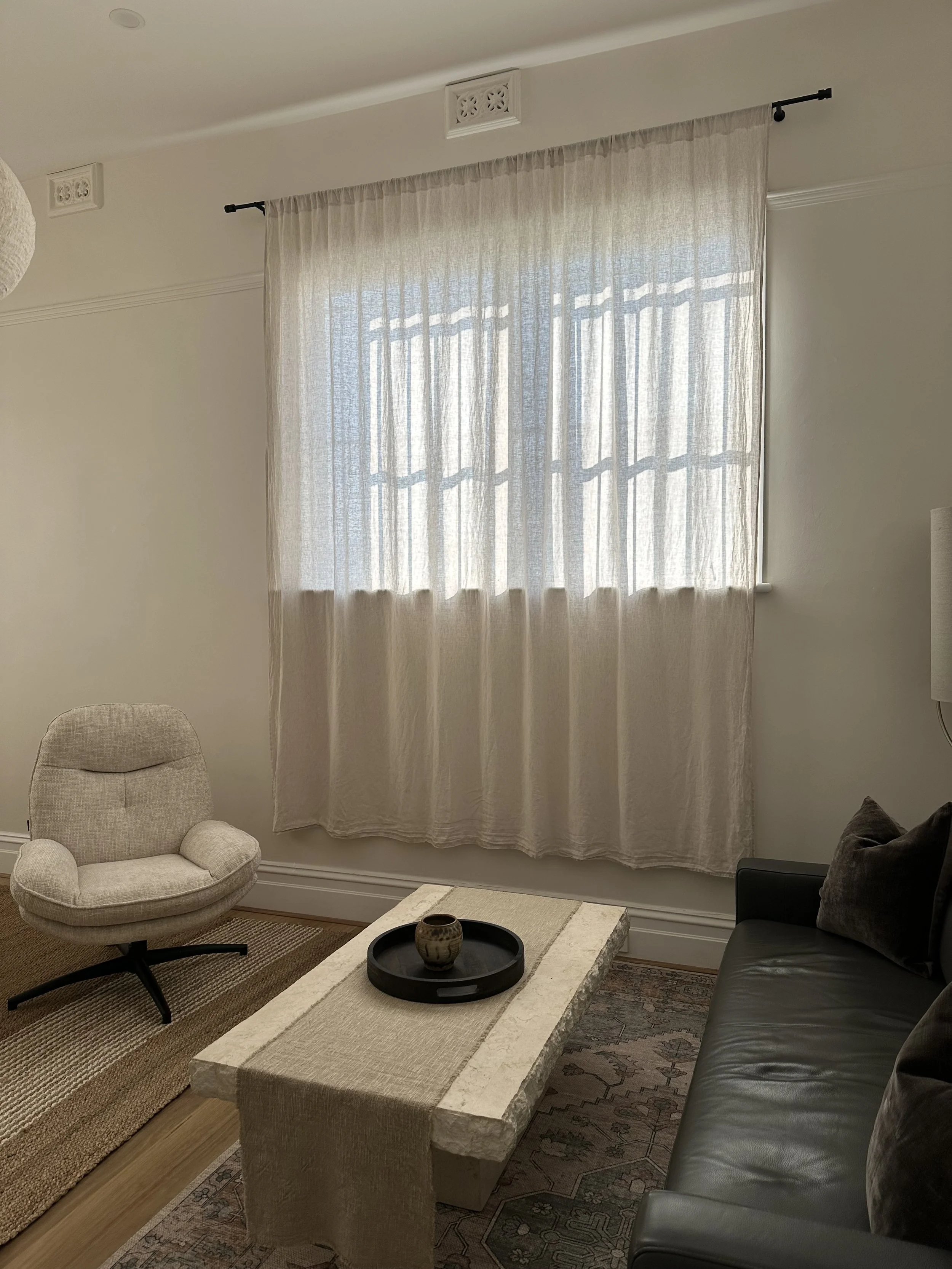 Psychotherapy consulting room with a beige armchair, a dark gray sofa with pillows, a coffee table with a decorative vase, a window with sheer curtains, and decorative ceiling vents.