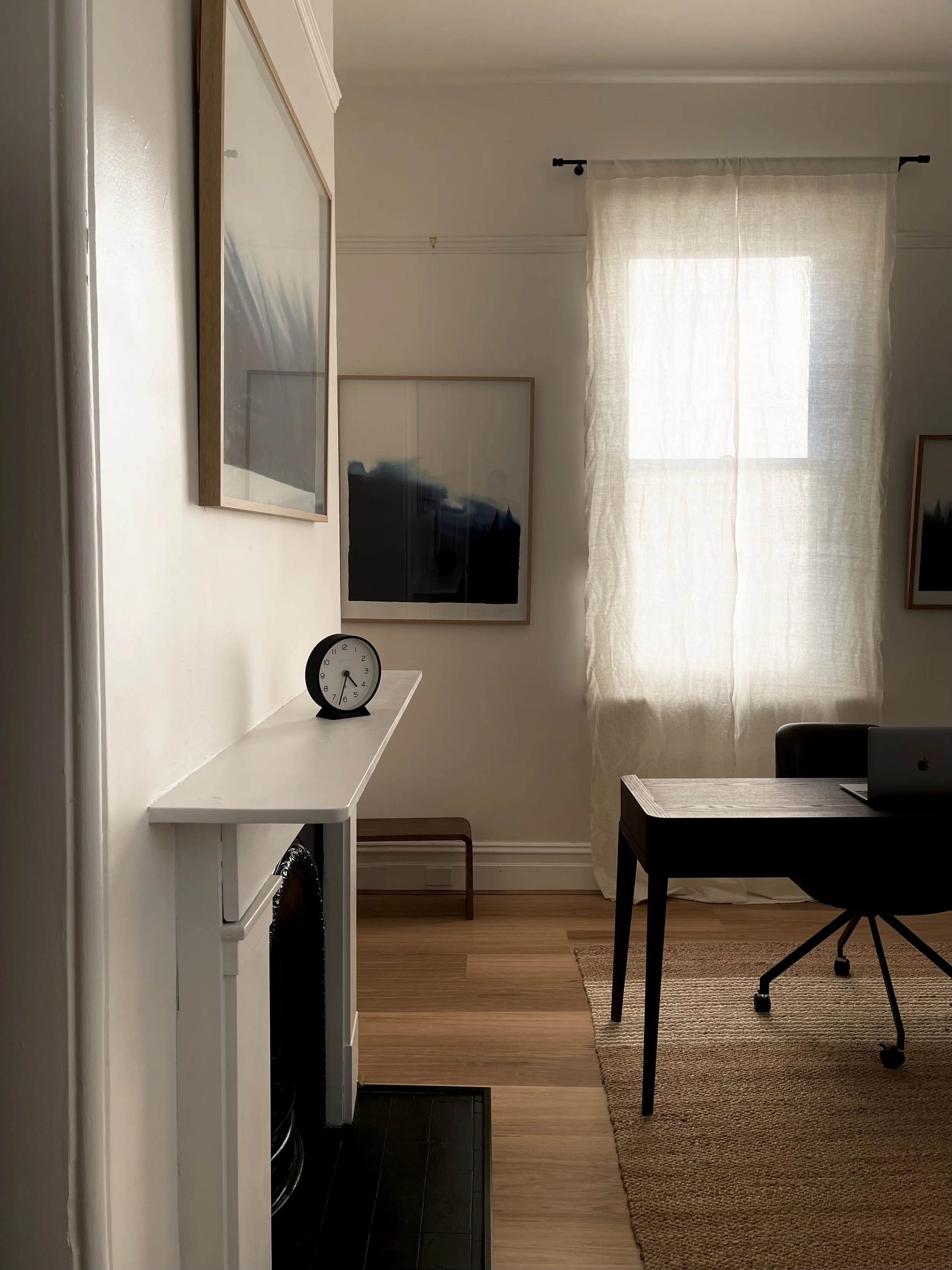An office desk in a psychotherapy consulting room with white walls, a window with light curtain, a black desk with a laptop, a white shelf with a clock, framed artwork, and a beige rug over wood flooring.