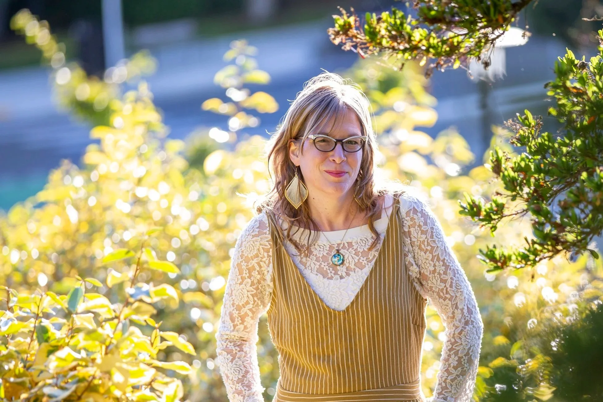 A woman with glasses and earrings smiling outdoors among yellow and green leaves, wearing a striped dress with lace sleeves and a pendant necklace.