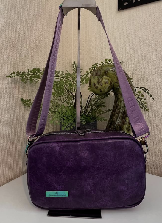 A purple leather shoulder bag with a teal tag, hanging on a stand against a beige wall, with a potted fern and a decorative green spiral-shaped sculpture in the background.