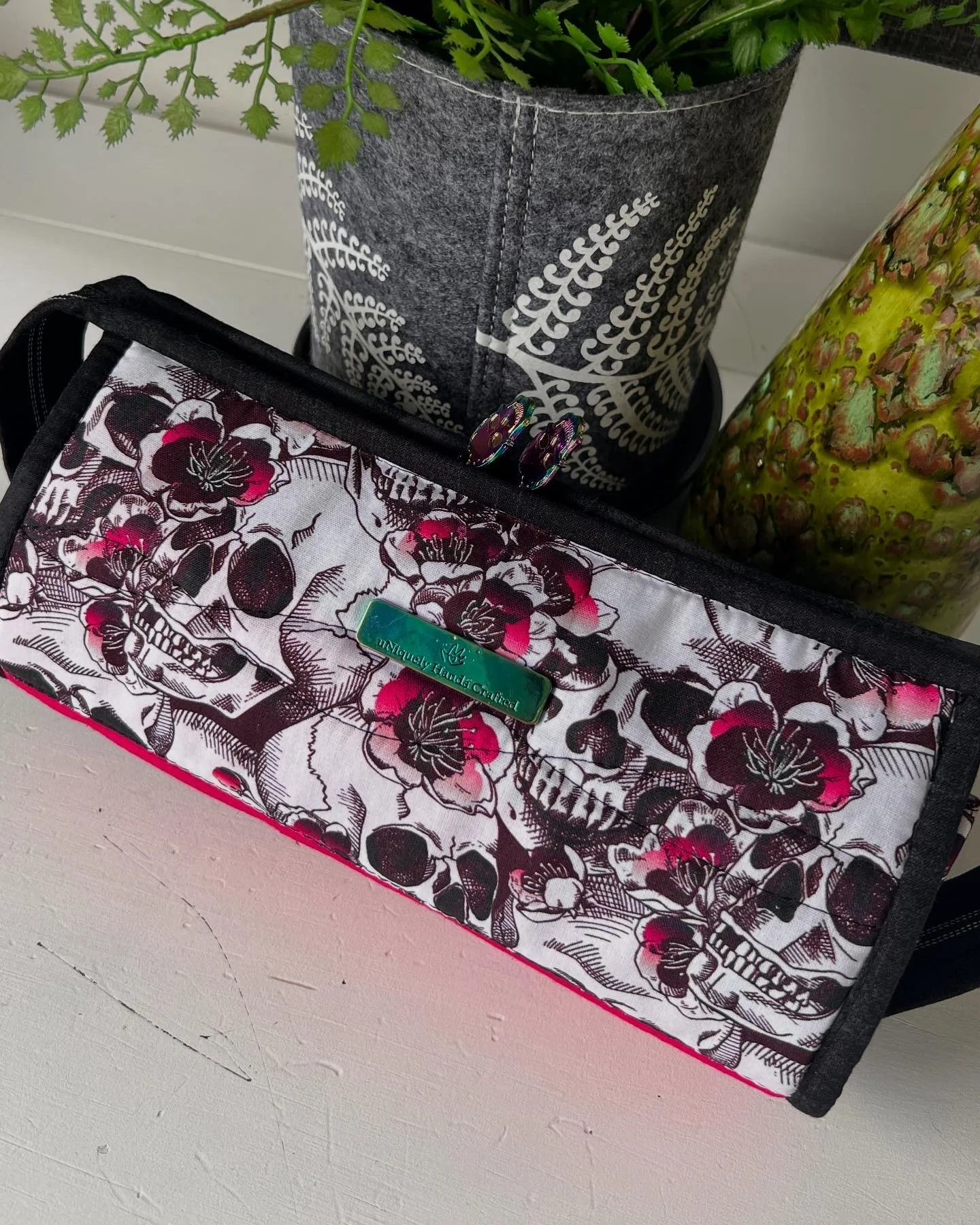 A black and pink floral fabric makeup or storage bag with a label that reads "Uniquely Hand Crafted." Inside the bag, there are two rings, and behind it, there are two potted plants, one in a gray felt pot with white decorative patterns and the other
