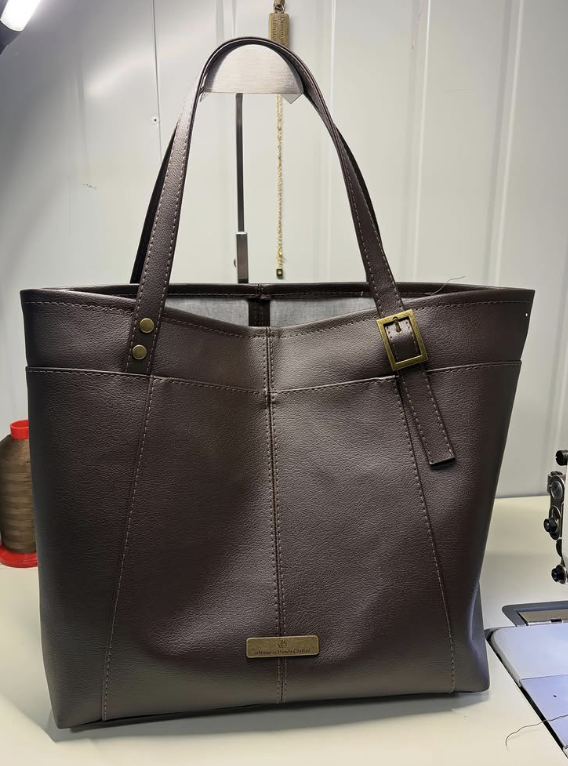 Brown leather tote bag with a gold buckle strap and a nameplate on the front