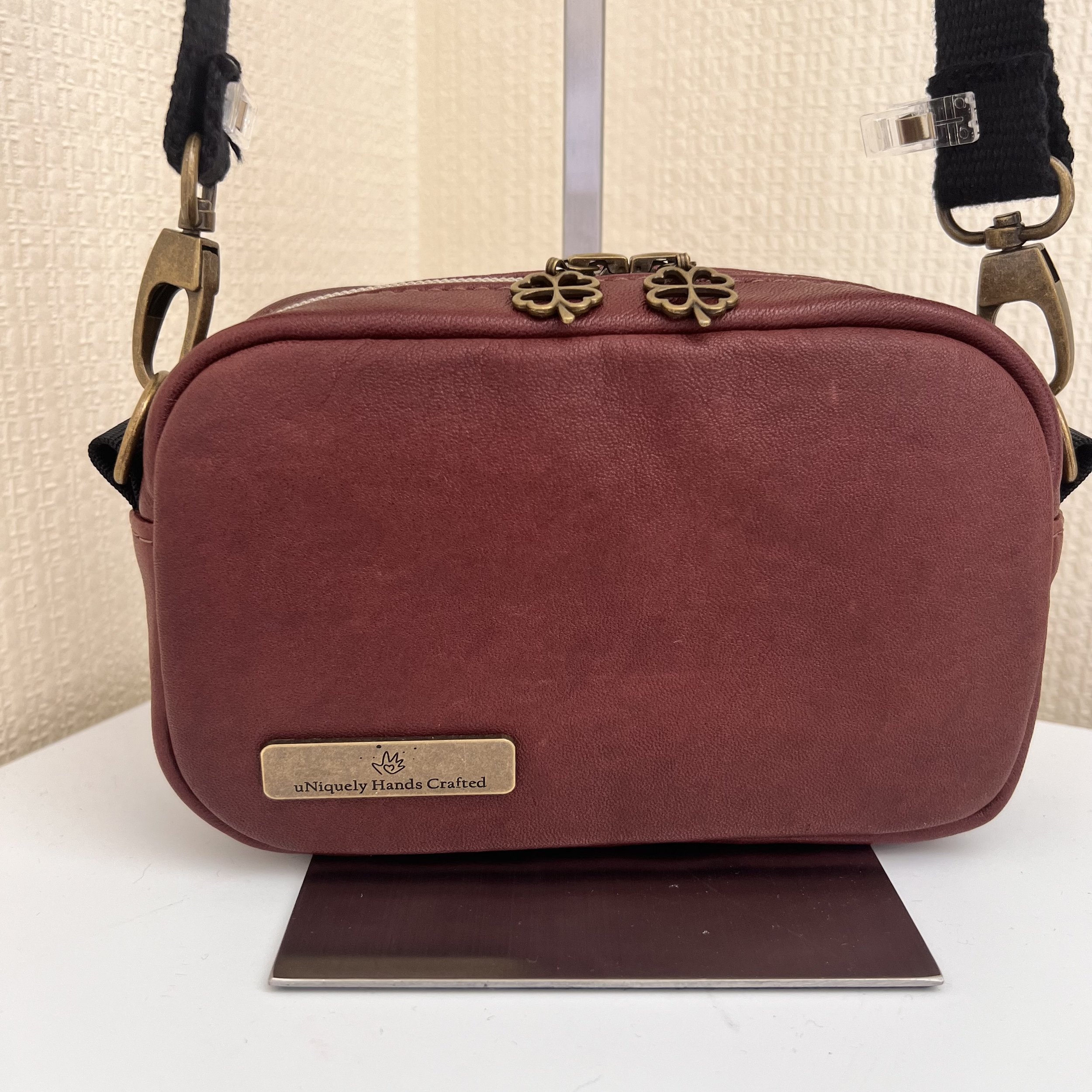 Red handbag with gold decorative charms on top, displayed on a stand with a metal plaque labeled 'uNiquely Hands Crafted' in front, against a light-colored textured wall background.