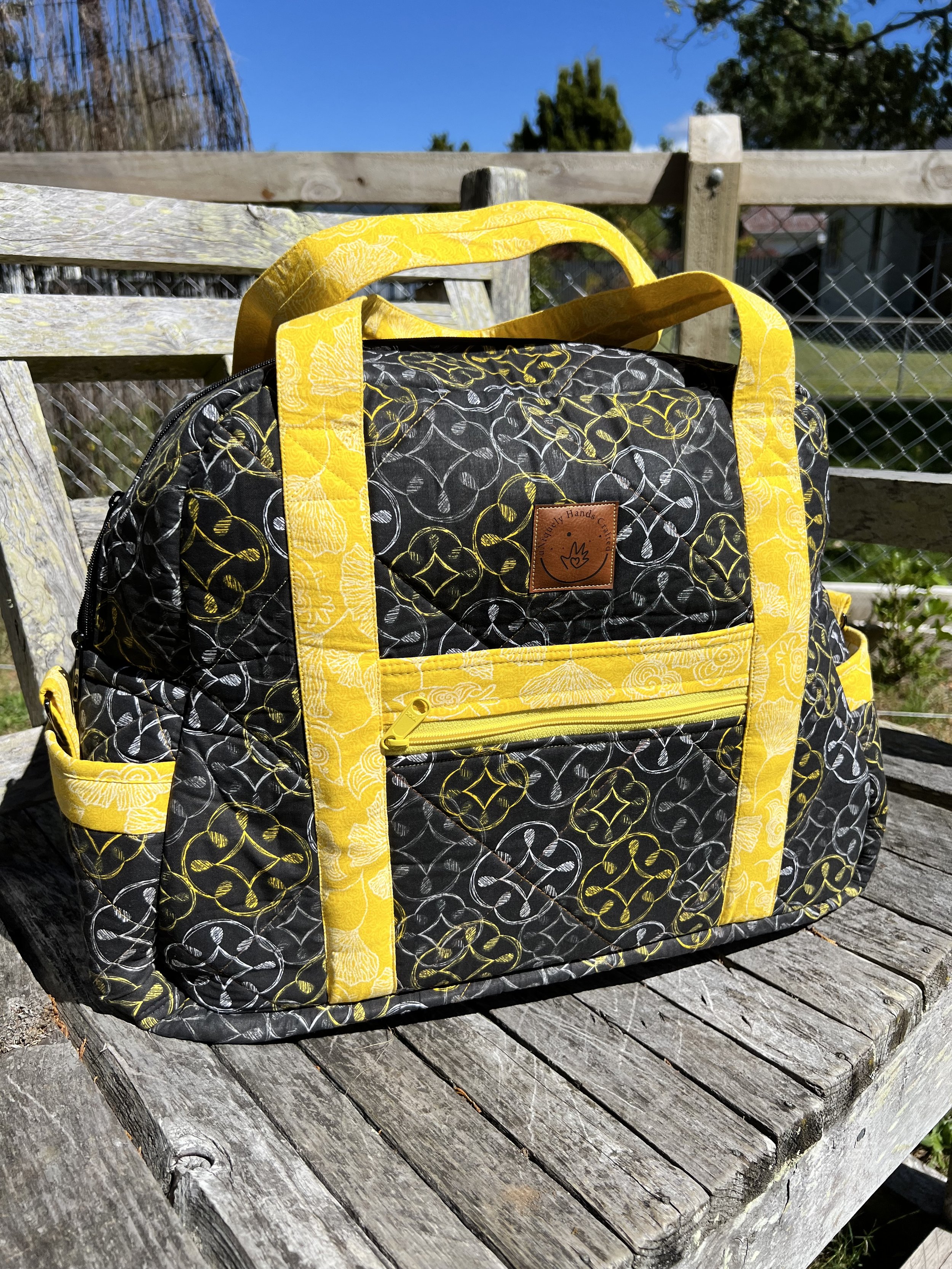 A black and yellow patterned bag with yellow handles and zippers placed on a weathered wooden surface outdoors, with a fence and trees in the background.