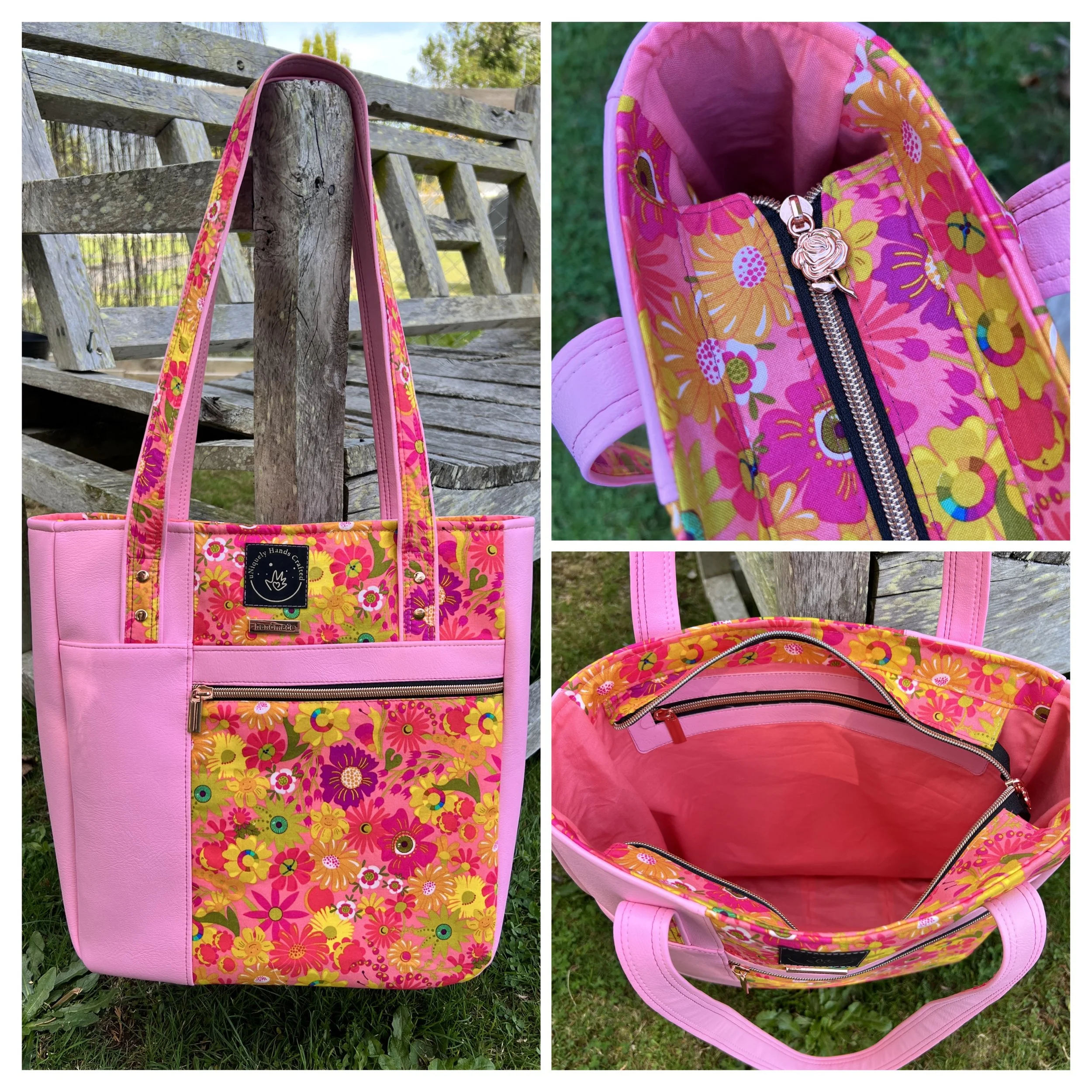 A pink floral tote bag hanging on a weathered wooden fence with green foliage in the background. The bag features bright pink, yellow, and orange flower patterns, a black label with a white logo, and a zipper pocket on the front.