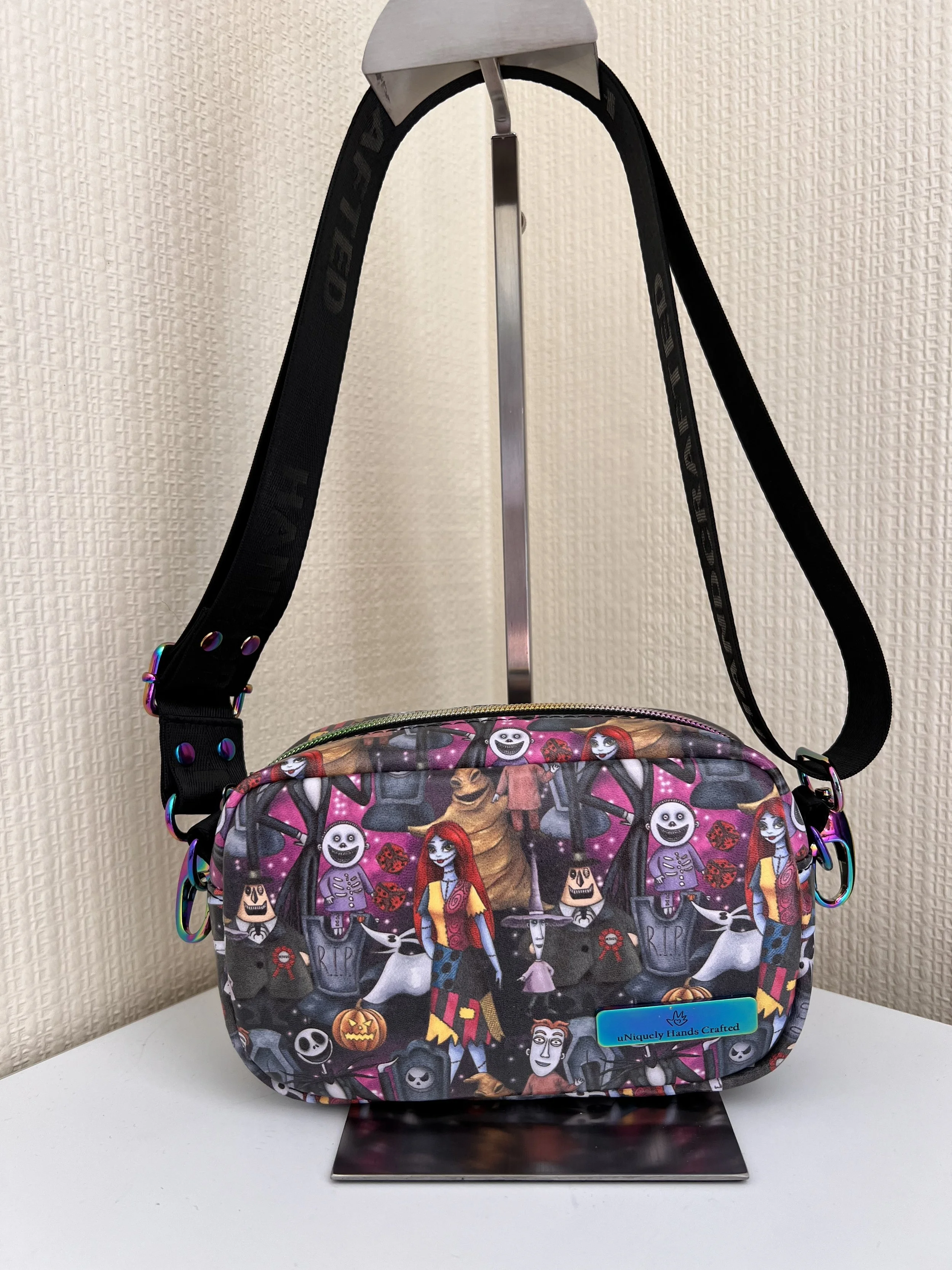 A Halloween-themed backpack with various spooky and playful characters, including skeletons, vampires, and pumpkins, on a white surface against a textured beige wall.