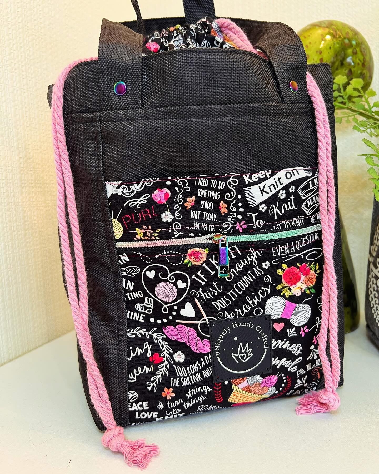A black fabric tote bag with pink braided rope handles, decorated with a colorful fabric patch featuring knitting motifs and words related to knitting, placed on a white surface near a beige wall with a plant and a small green and yellow fruit in the