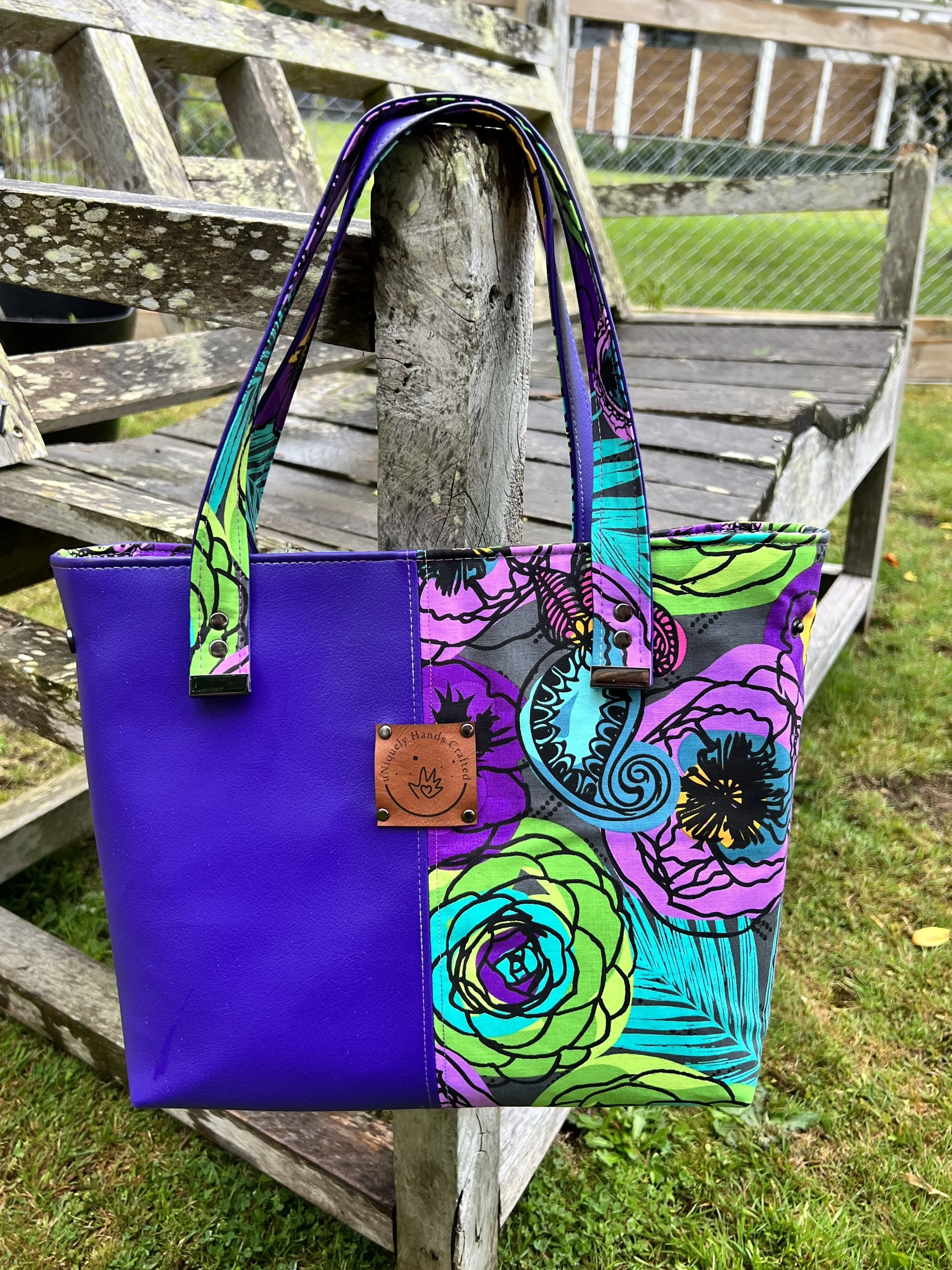Colorful floral tote bag with purple, green, and black patterns hangs on a wooden fence post outdoors.