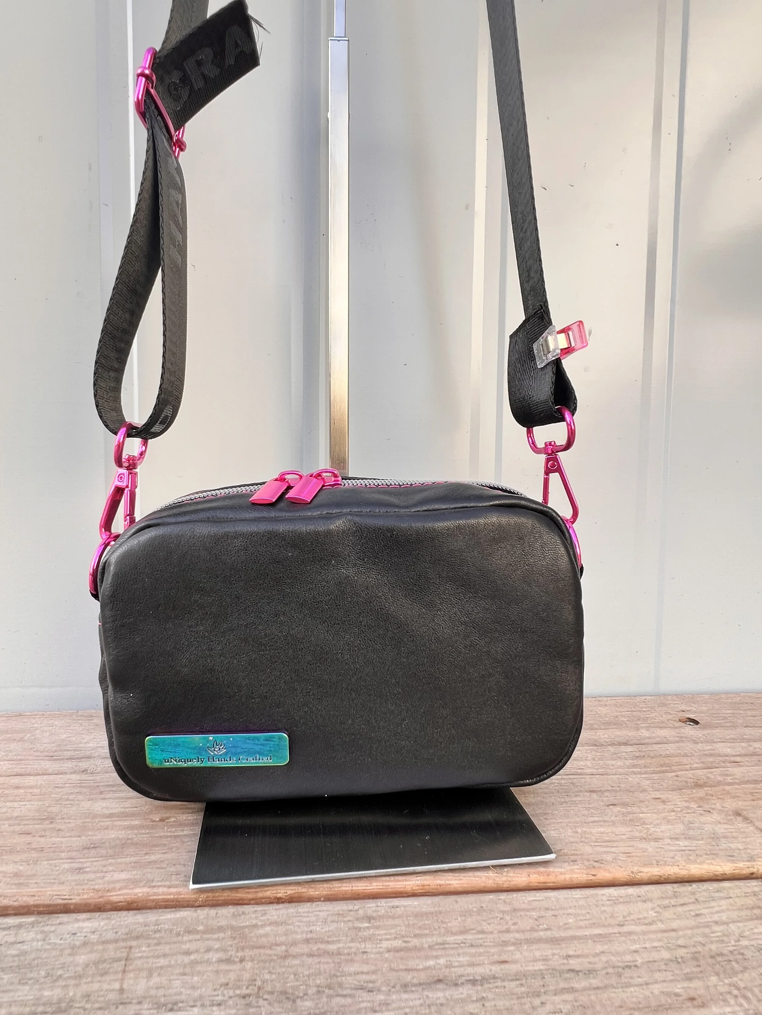 Black handbag with pink accents on a small scale model swing set, placed on a black rectangular base on a wooden surface.