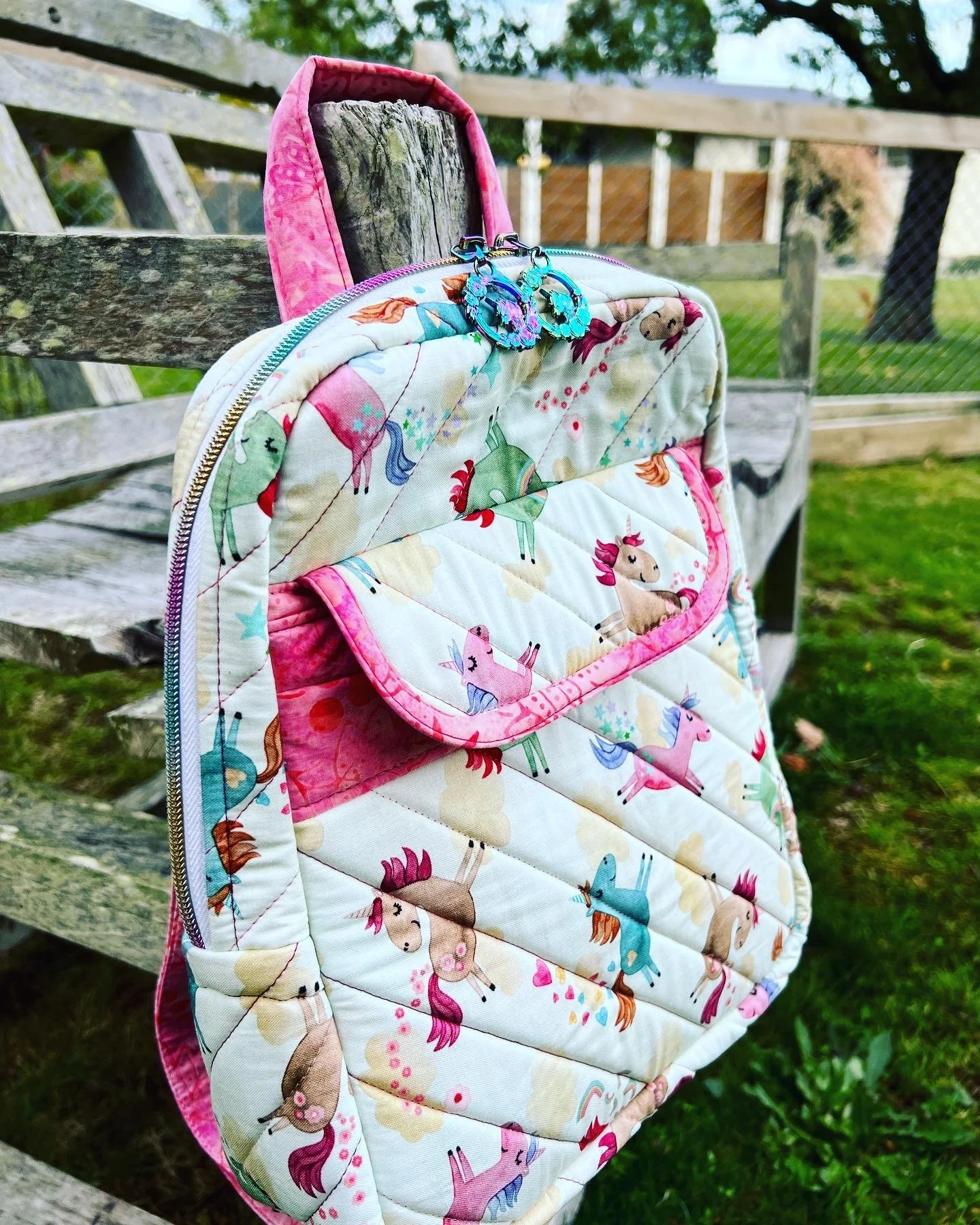 A colorful quilted backpack with a unicorn pattern hanging on a weathered wooden fence in an outdoor setting with grass and trees in the background.