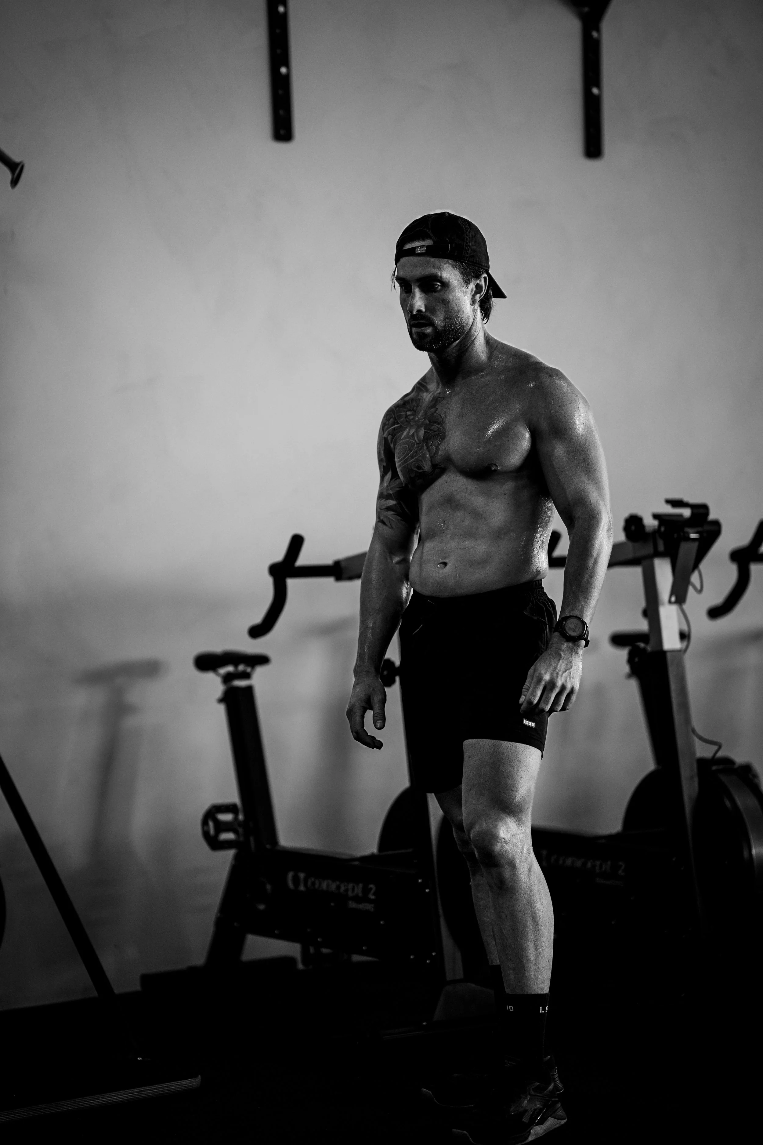A shirtless man wearing a backward cap and shorts, standing in a gym with exercise equipment in the background.