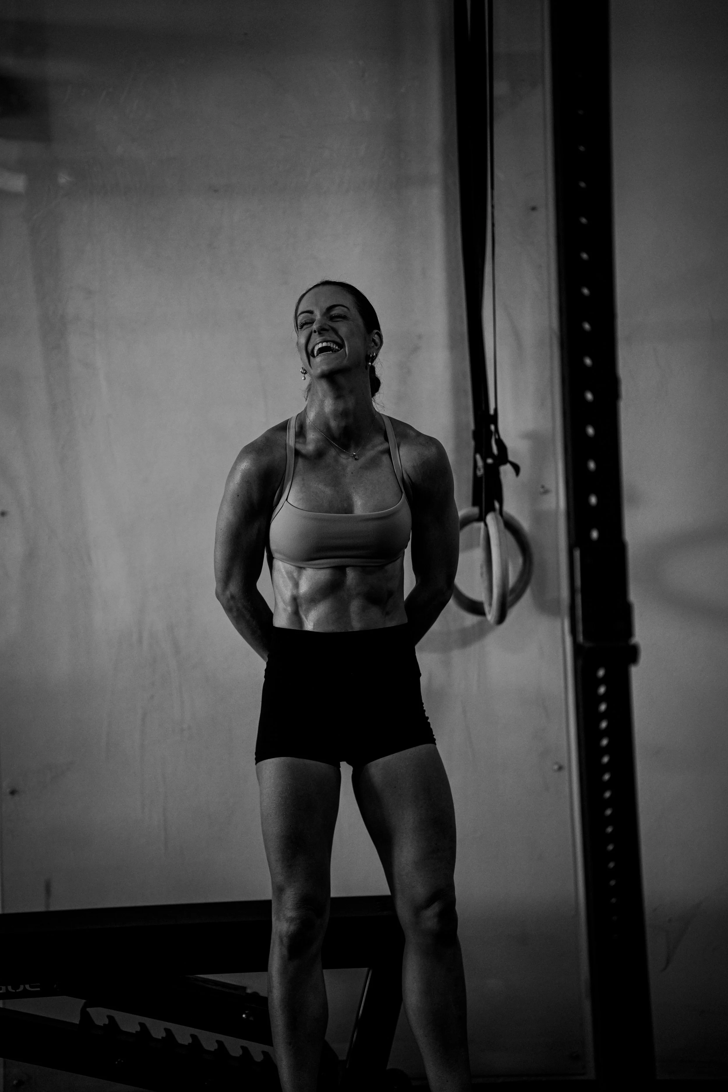 A woman in athletic attire smiling and standing in a gym. She is wearing a sports bra and shorts, with fitness equipment like rings and a pull-up bar visible in the background.