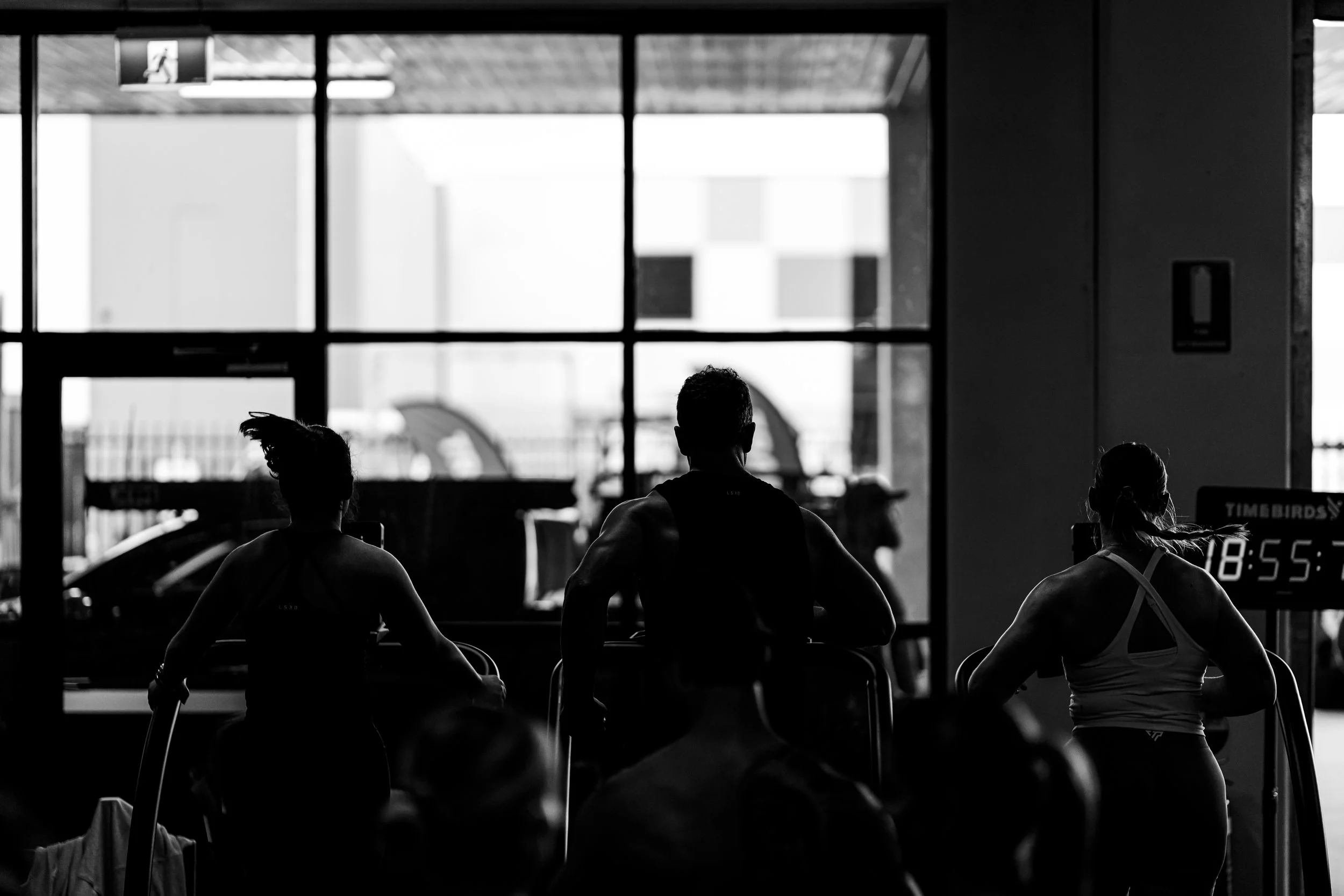 Silhouettes of people running on a treadmill in a gym with large windows and a digital clock showing 18:55.