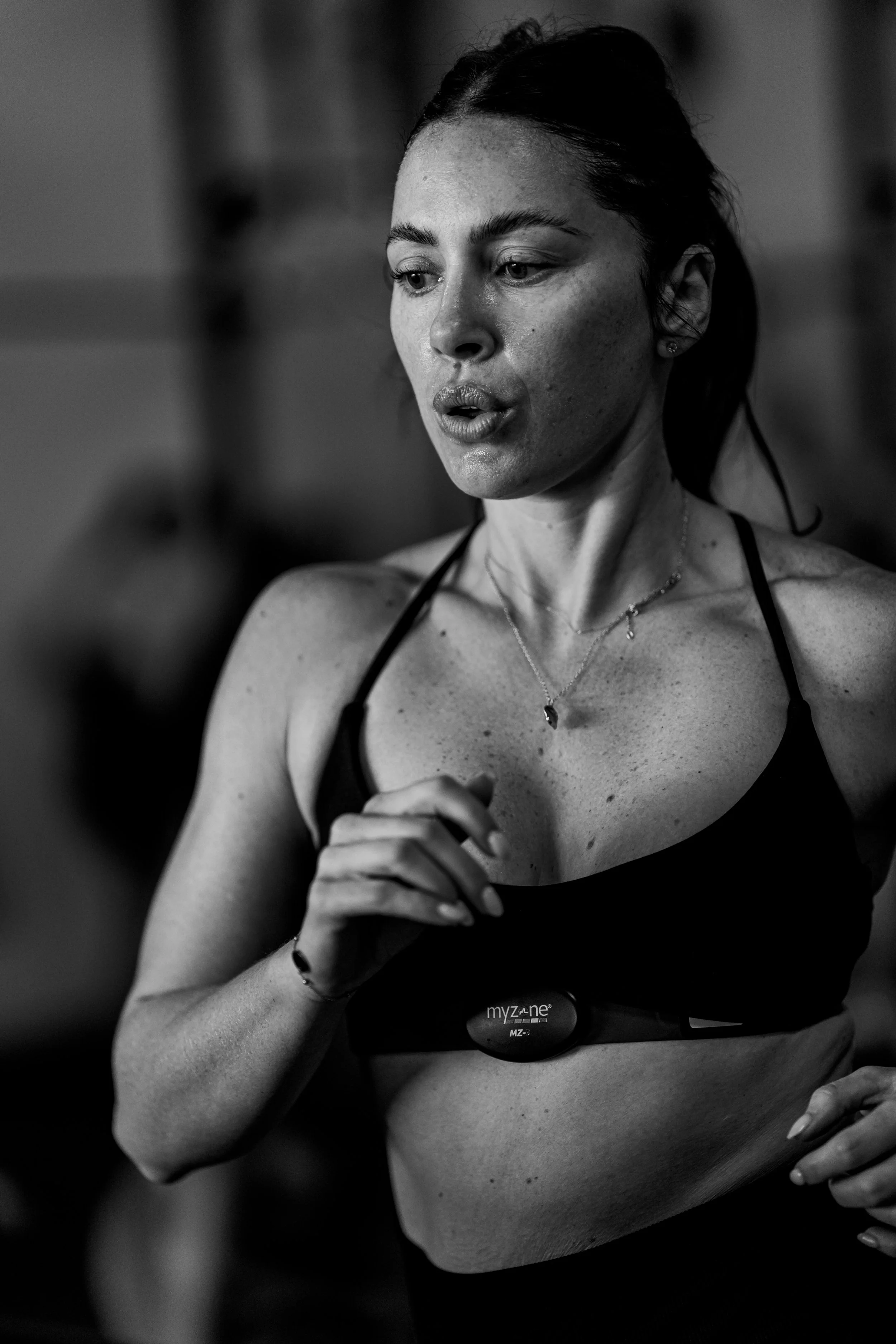 A black and white photo of a woman with water droplets on her face and shoulders, wearing a sports bra and jewelry, holding her hand close to her chest.