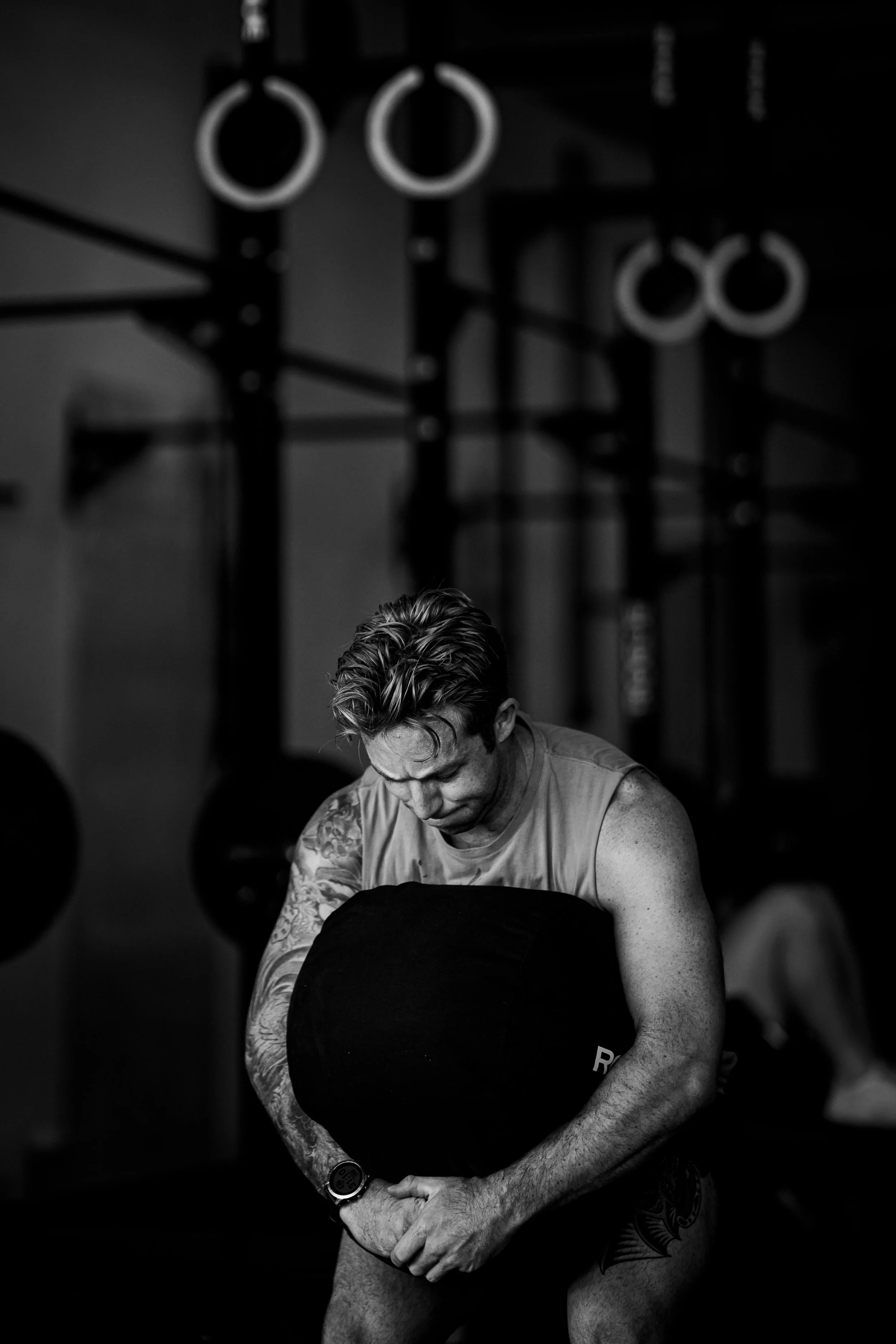 A man with tattoos on his arms and chest, wearing a sleeveless shirt, is adjusting his shorts in a gym with black and white gymnastic rings hanging from the ceiling.