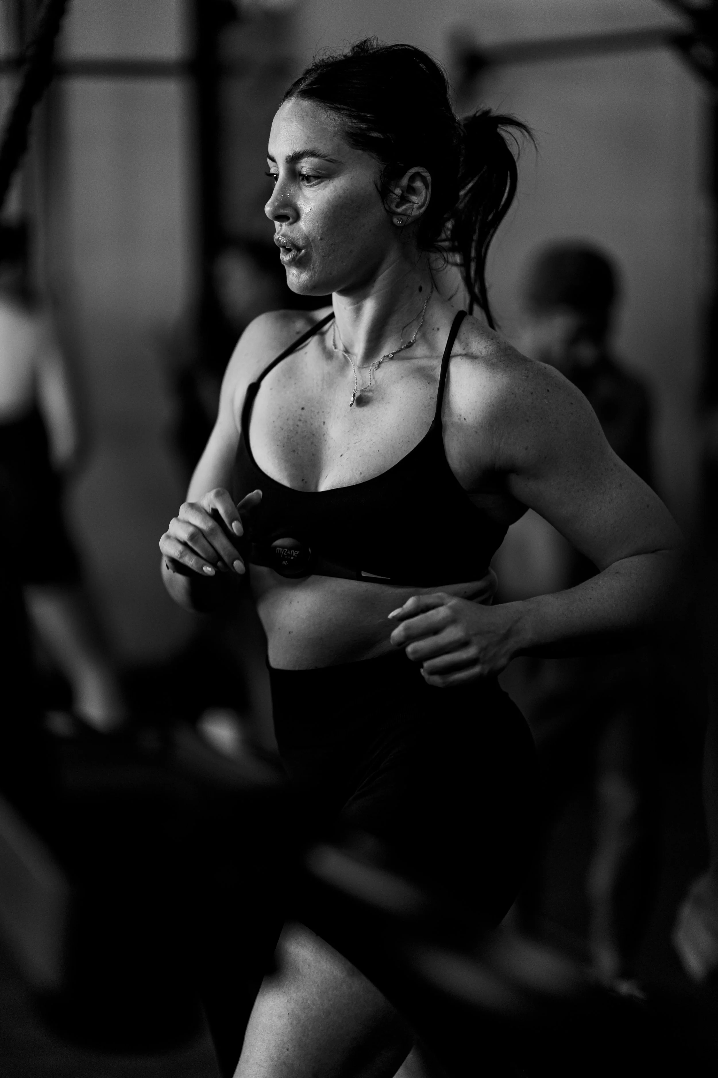 Black and white photo of a woman running in a gym, focused and determined, wearing a sports bra and a necklace.