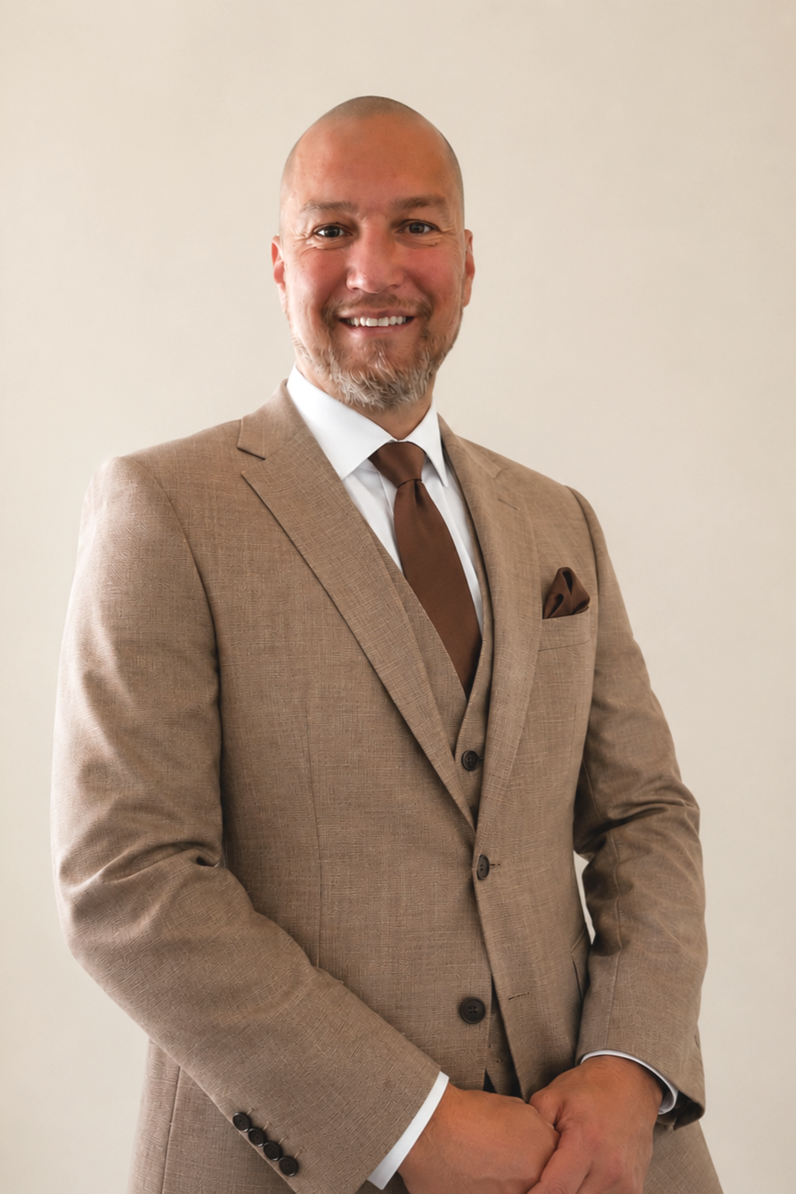 A man smiling in a beige suit with a brown tie and pocket square, standing against a plain light-colored wall.
