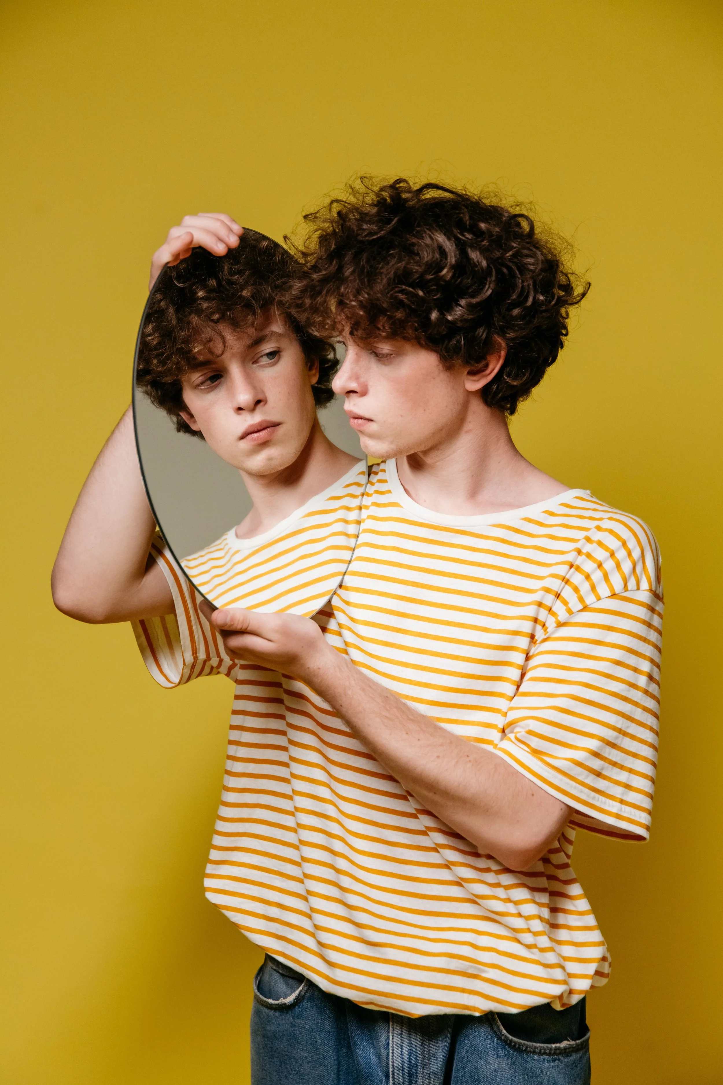 Young man with curly brown hair in a yellow and white striped t-shirt holding a mirror, looking at his reflection, standing against a yellow background.
