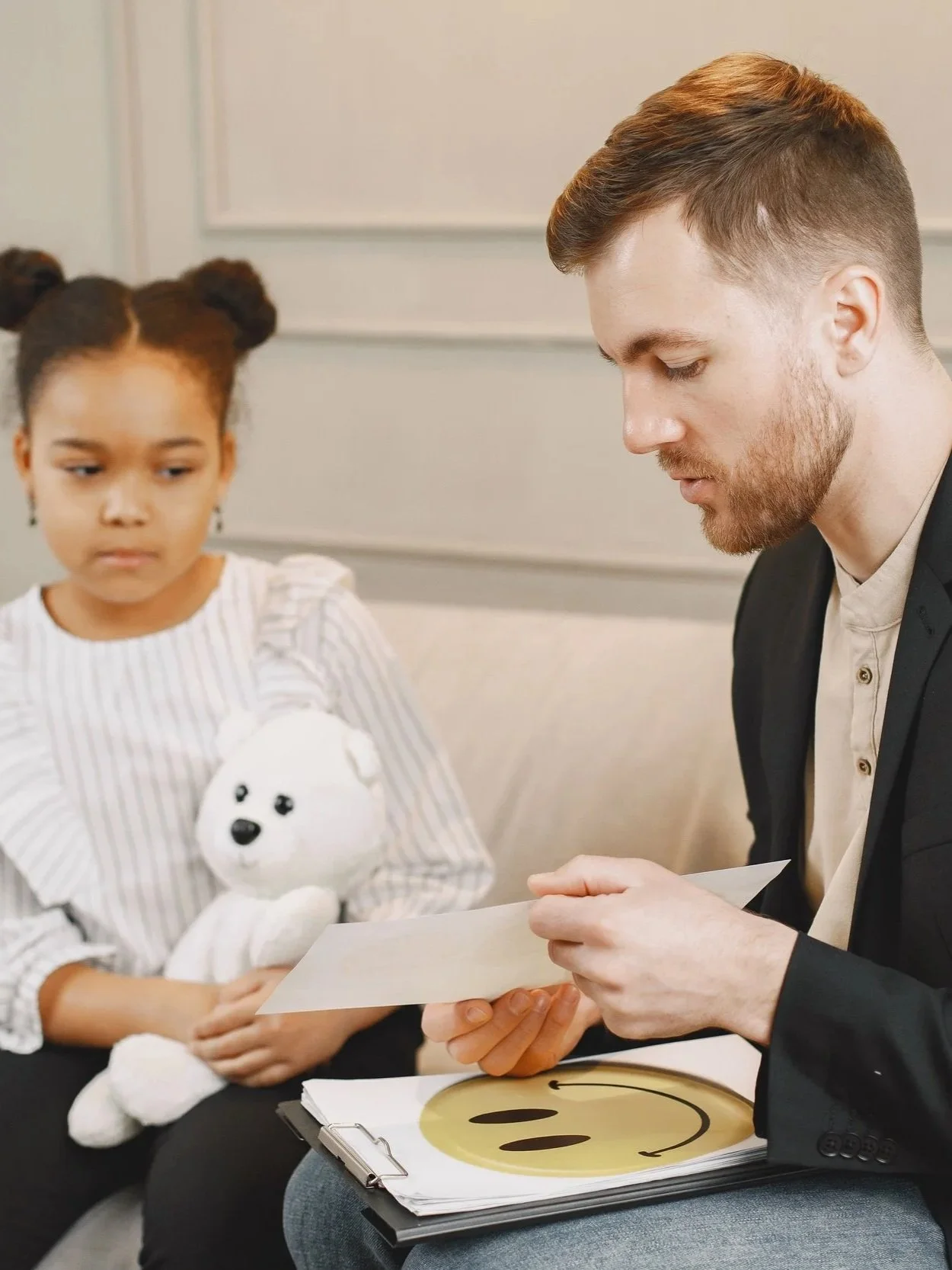 A young girl with braided hair holding a stuffed bear, sitting next to a man in a blazer looking at a piece of paper, with a folder displaying a smiley face icon on her lap.