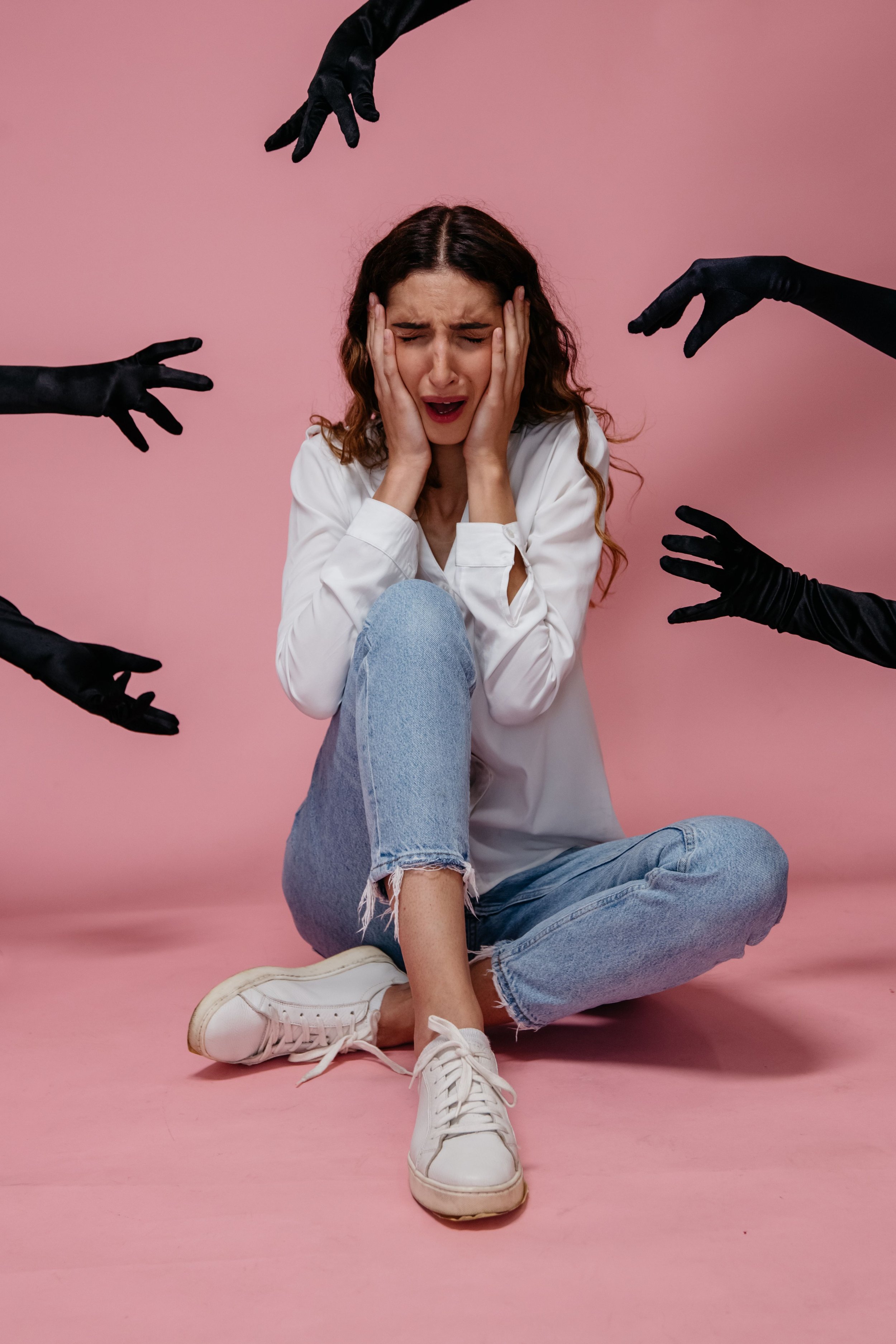 A young woman wearing a white shirt and light blue jeans sitting on the floor with a pink background. She appears distressed with her hands on her face. Surrounding her are four black-gloved hands reaching towards her.