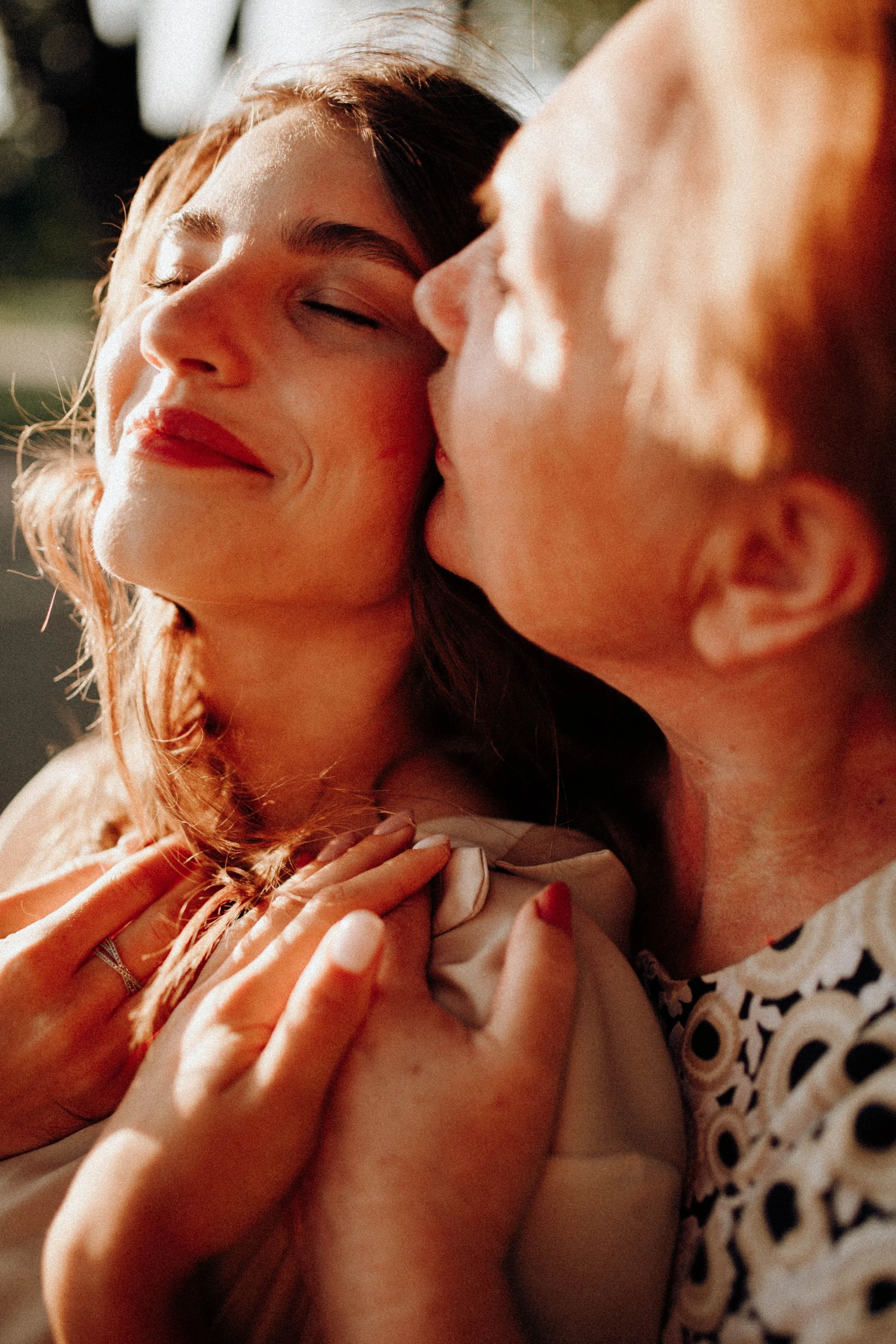 A woman with brown hair and makeup, smiling with eyes closed, being kissed on the cheek and forehead by a man with light hair, both in close embrace and holding hands.