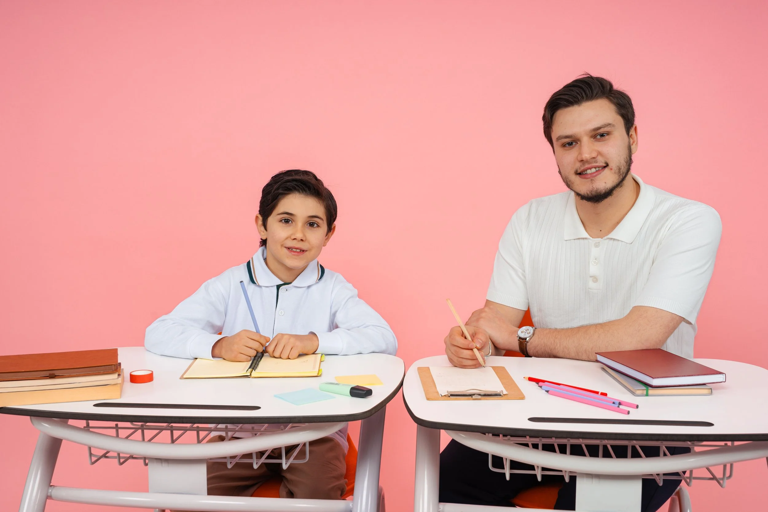 A young boy and a young man are sitting at desks with notebooks and pens, against a pink background. They are smiling and looking at the camera, suggesting a classroom or study setting.