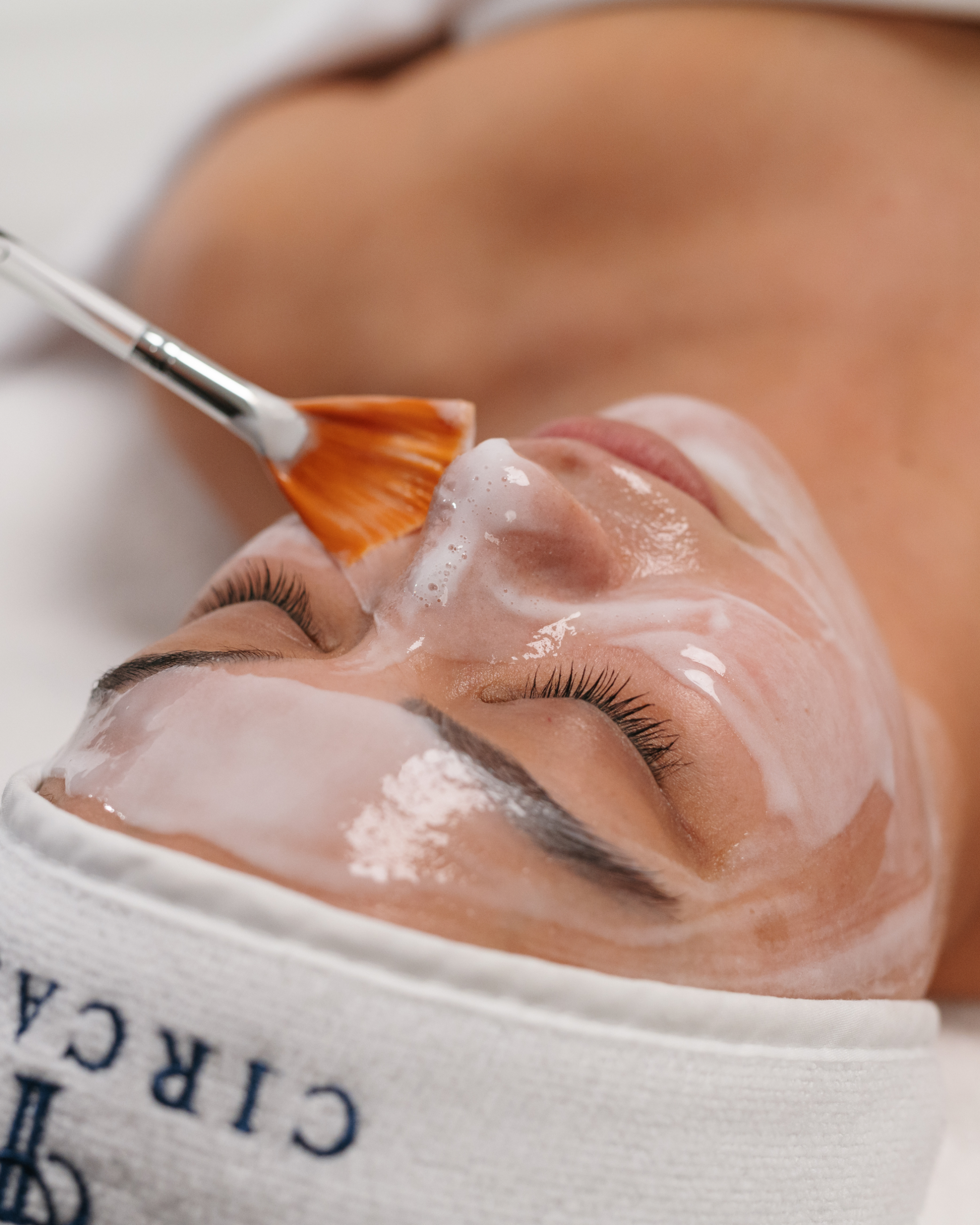 Close-up of a woman receiving a facial treatment with a brush applying cream or mask to her face, her eyes closed, lying on a treatment bed.
