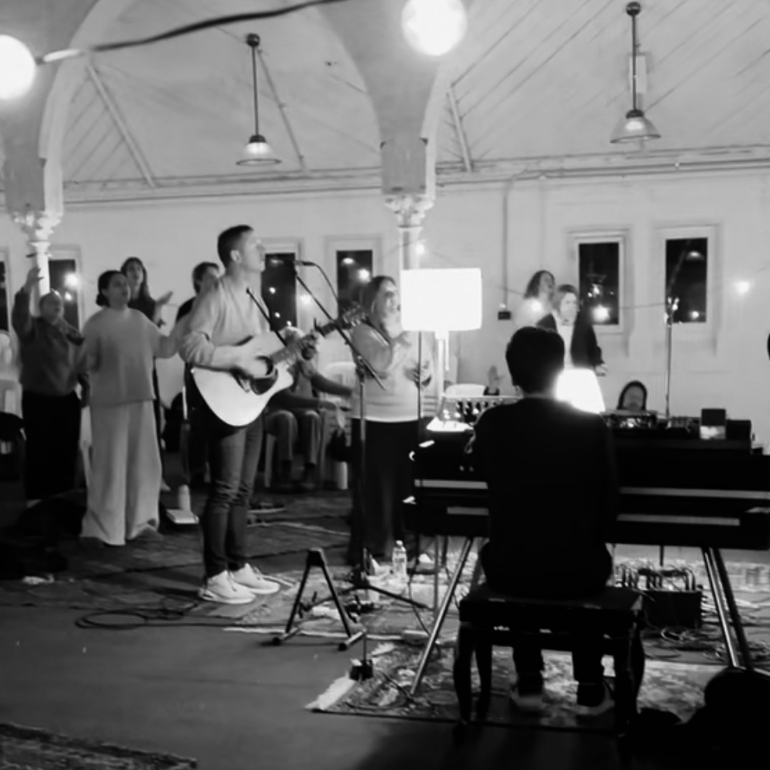 Black and white photo of a group of people performing music in a room with hanging pendant lights and large windows. A person is playing an acoustic guitar and singing into a microphone, while others are standing or sitting around, possibly singing or playing instruments.