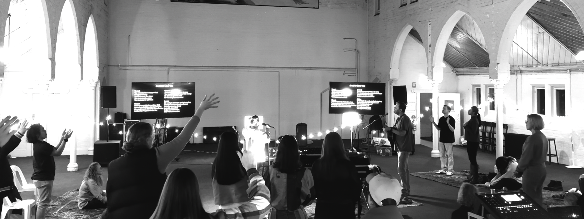 A group of people in a church or community space gathered for a service or event, with some raising their hands, a person speaking or singing into a microphone, and a large screen displaying text.