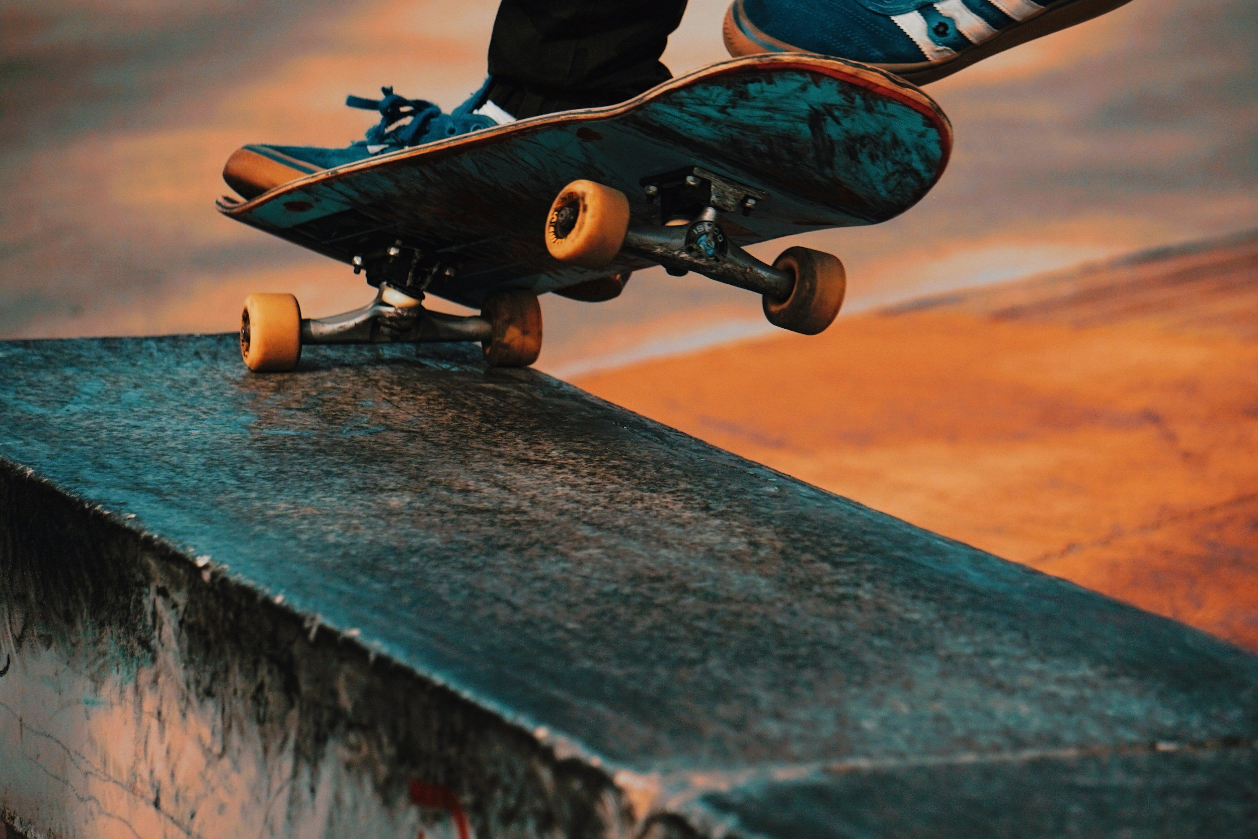 Close-up of a skateboard on a skate ramp at sunset, showing the skateboard's underside and wheels, with a person's feet wearing striped shoes in the background.