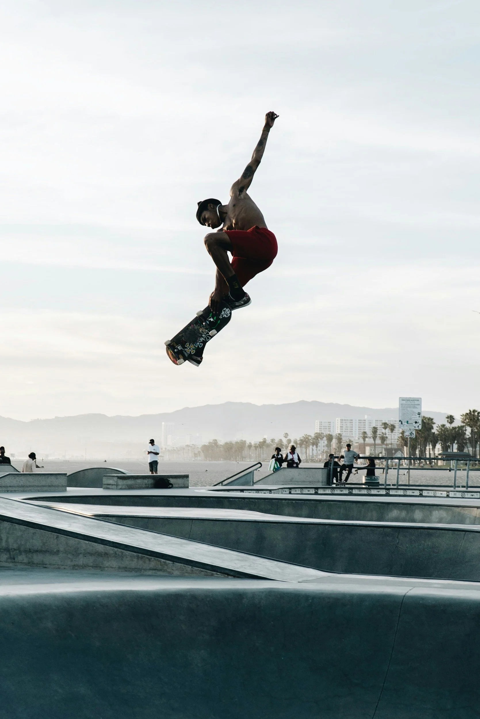 A person skateboarding in a skate park near the beach, performing an aerial trick in the air, with a group of people watching in the background, under a cloudy sky.