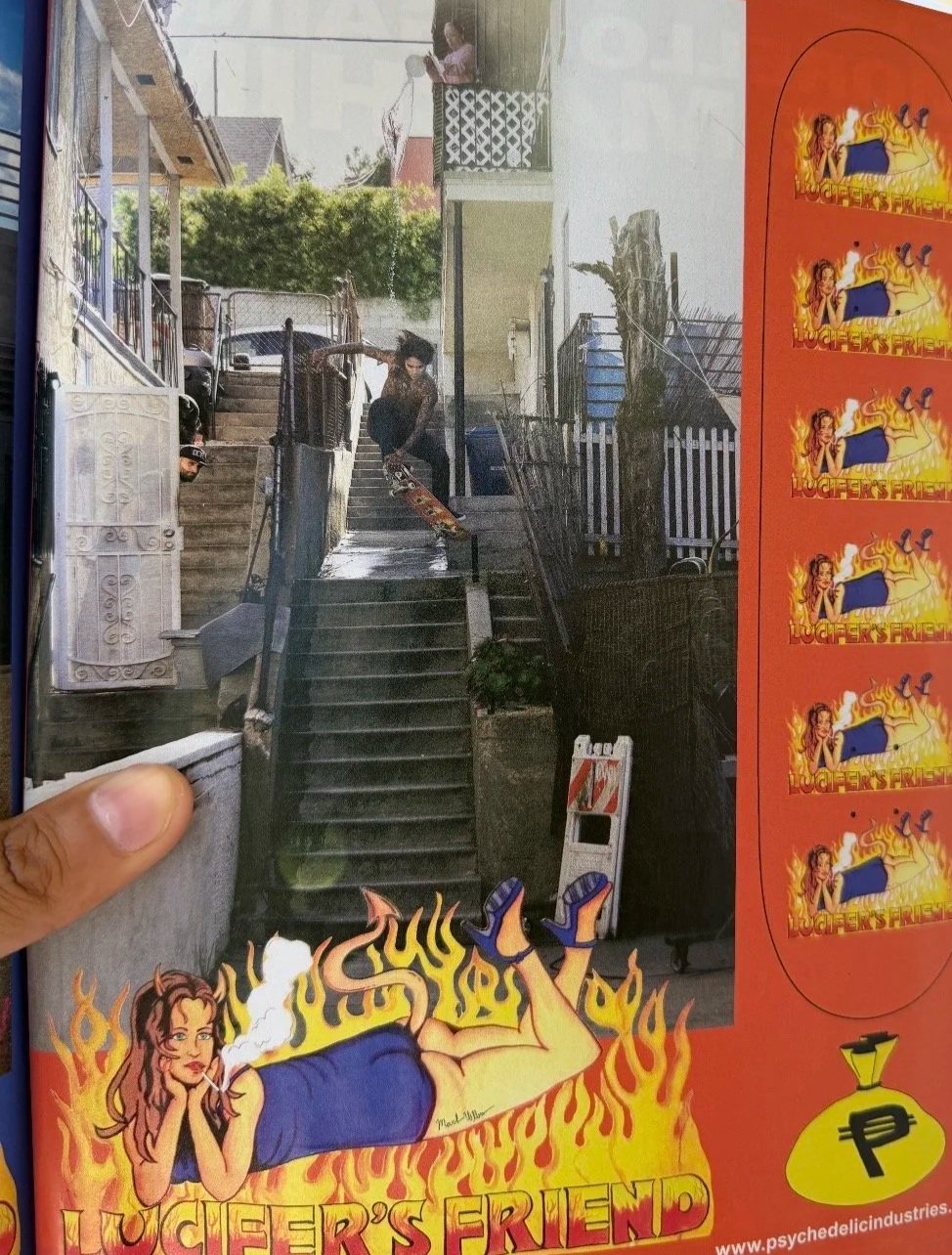 Photograph of a steep outdoor staircase in a residential neighborhood, with skateboarders performing tricks, a person sitting on a balcony, and promotional stickers for Lucifer's Friend band on the right side.