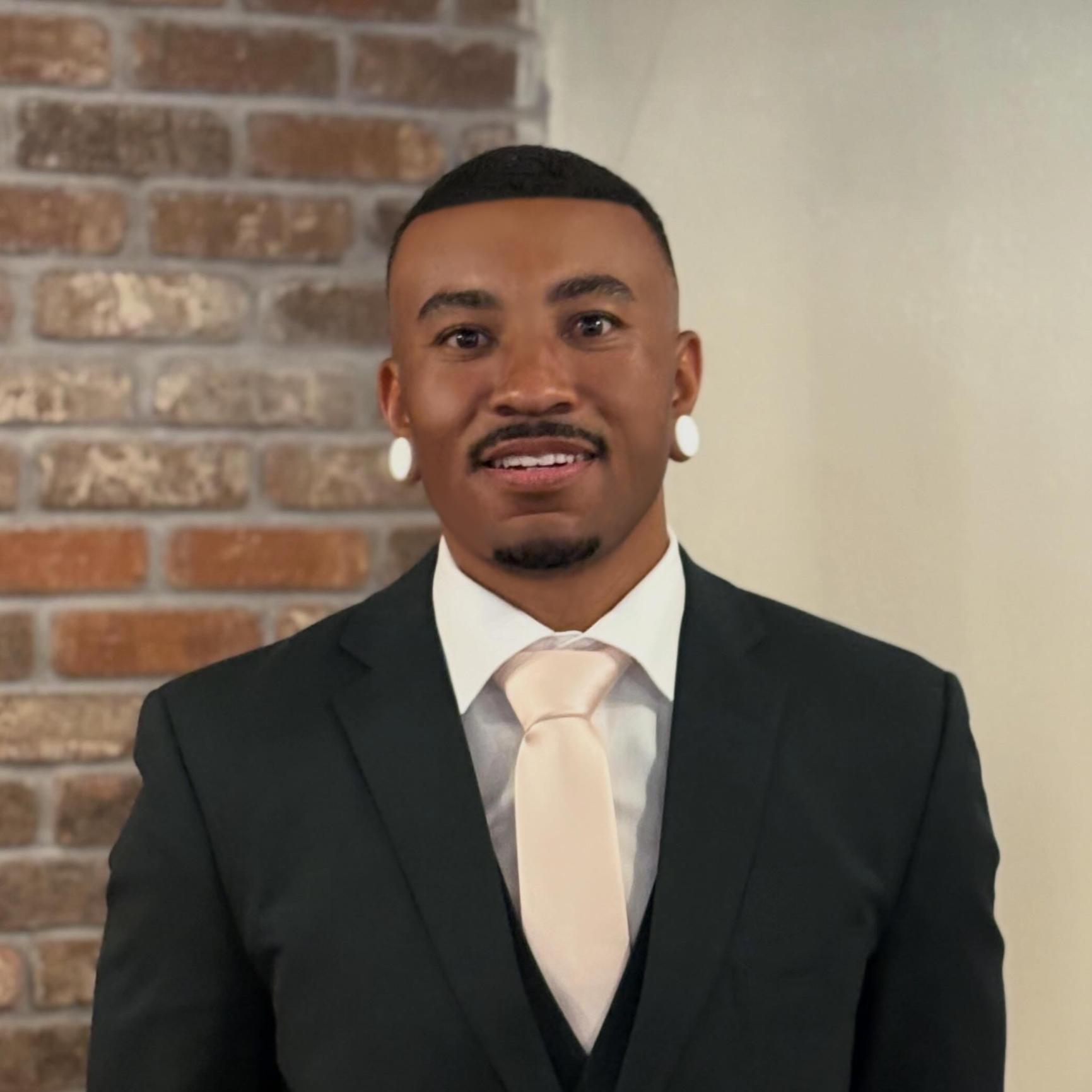 A man in a tuxedo with a white shirt and a light-colored tie, standing in front of a brick wall and a plain light-colored wall, smiling at the camera.