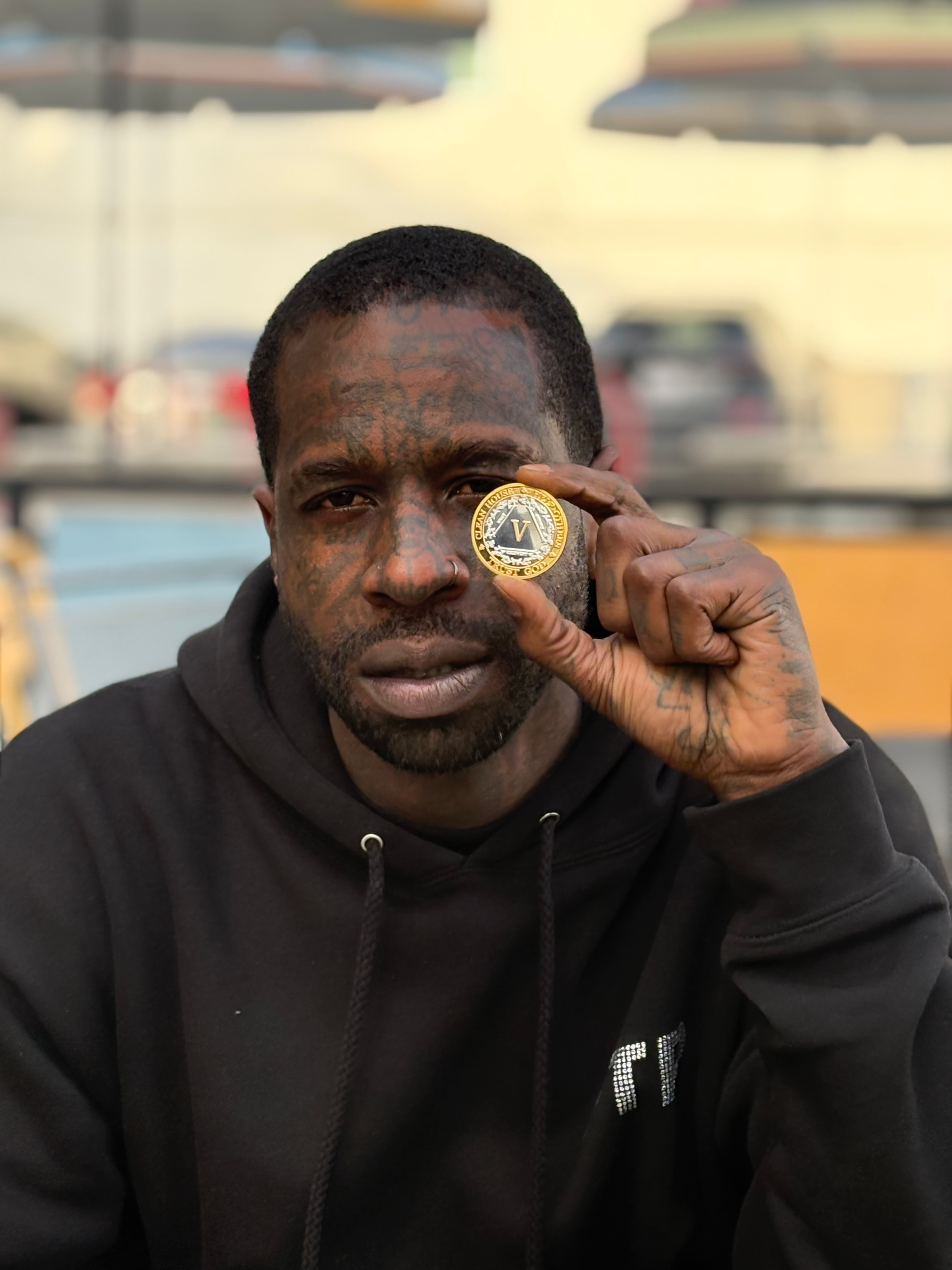 A man with tattoos on his face and hand, holding a coin with a 'V' on it over his eye, wearing a black hoodie, with a blurred parking lot in the background.
