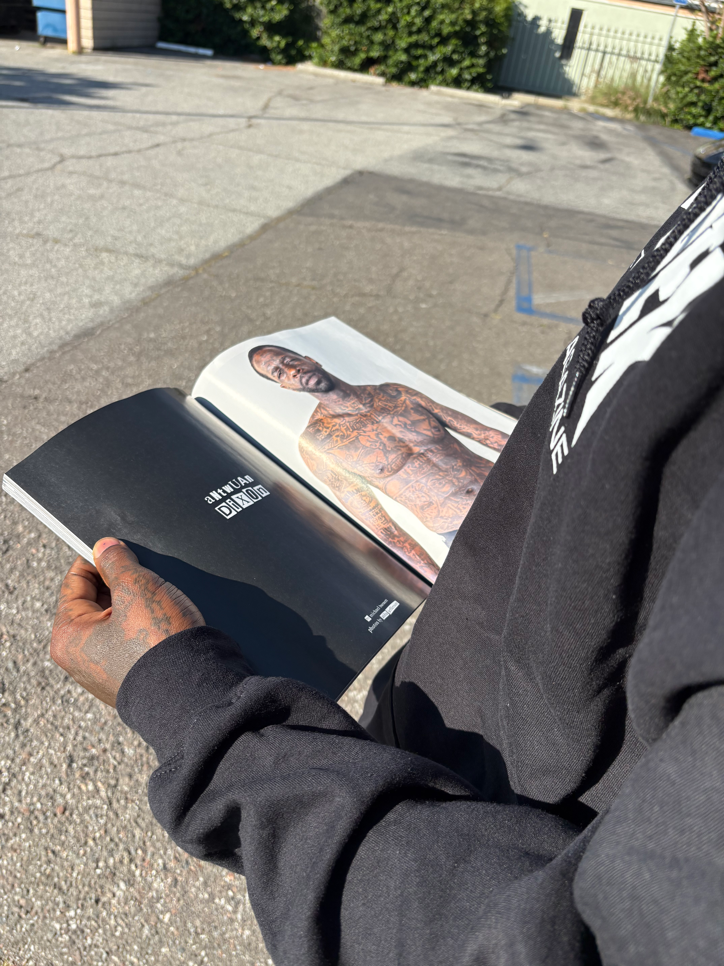A person sitting outdoors reading a magazine with a picture of a heavily tattooed man on the cover. The person's hand and part of their black clothing are visible, and they are seated on a black chair or stool. The background shows a parking lot with cracks in the pavement, some bushes, and a fence.