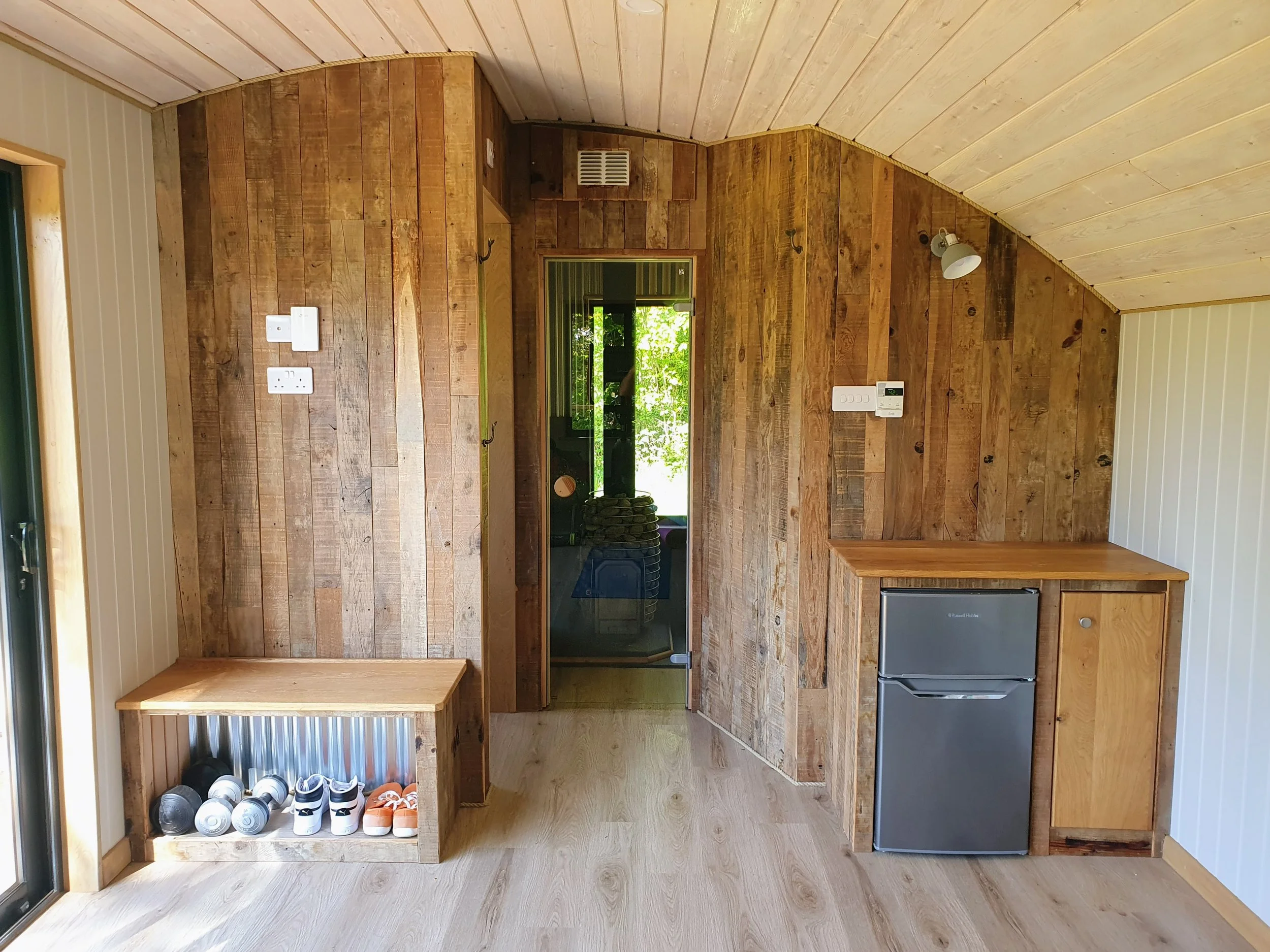 A small kitchen with wood-paneled walls and ceiling, featuring a window with outdoor greenery visible, a small compact refrigerator, a black oven, a wooden countertop, and a shoe rack with shoes on it.