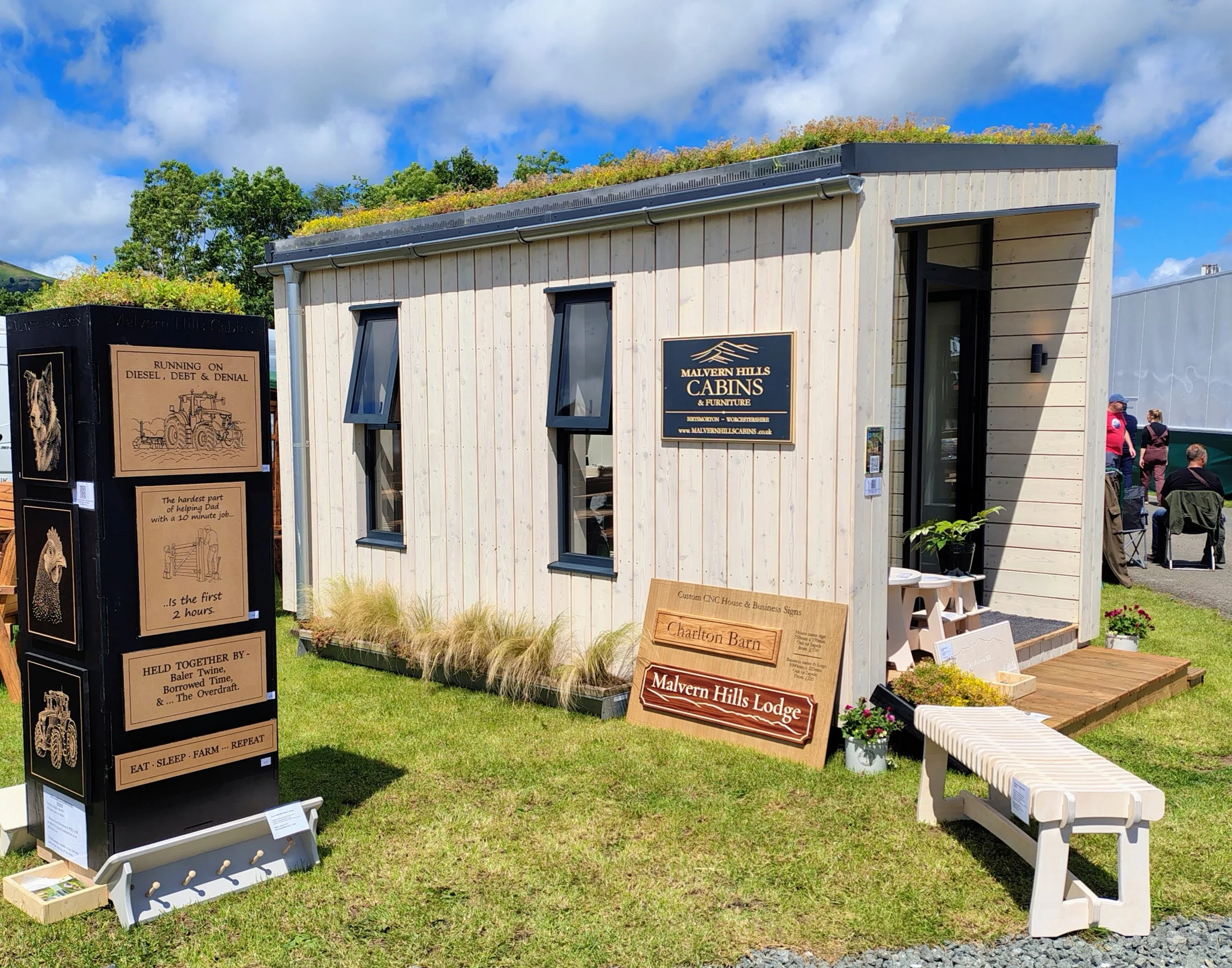 An outdoor display booth at a fair or event featuring wooden furniture, signs, and decorative items for sale. There are people in the background, a bench, and a building with a sign that reads "Malvern Hills Cabins & Furniture".