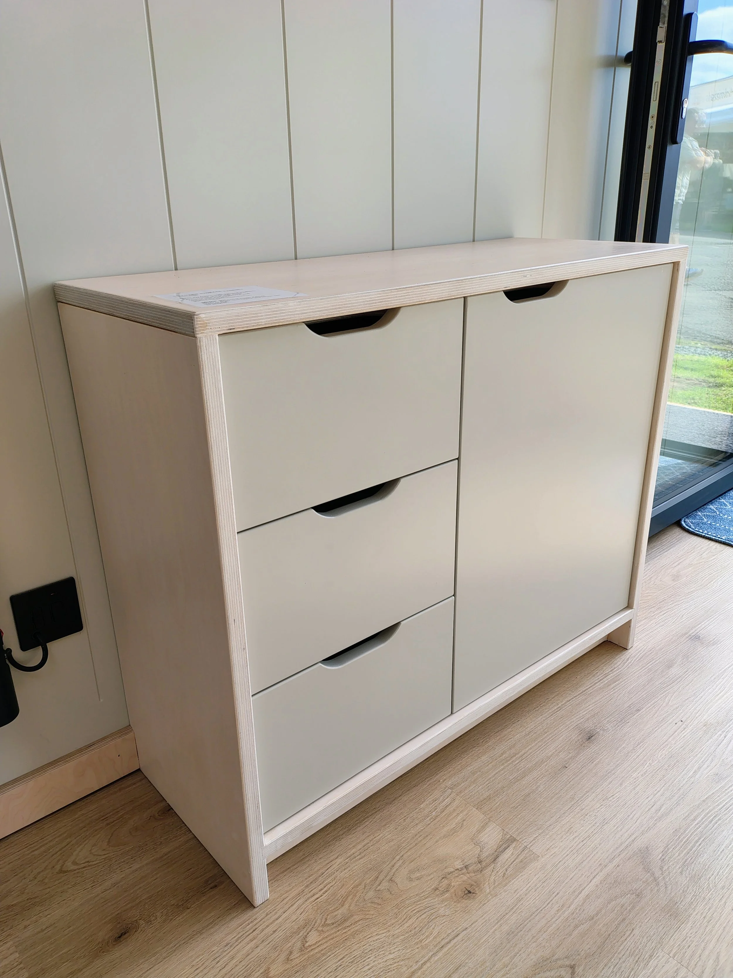 White wooden cabinet with multiple drawers, positioned near a wall with vertical paneling and a glass door, on a light wood floor.