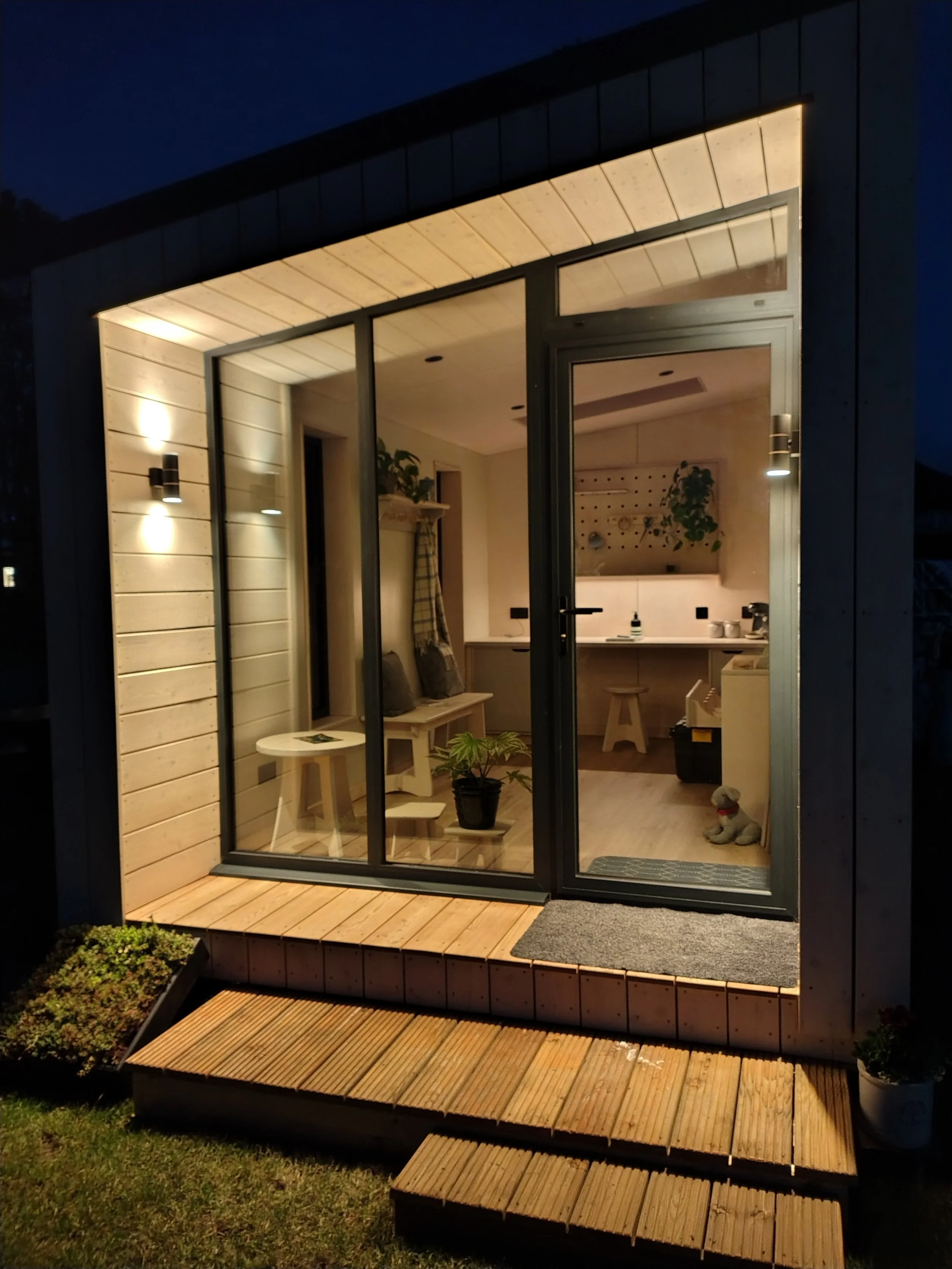 A small modern house with large glass windows and sliding doors, illuminated from inside, showing a cozy interior with plants, furniture, and decorations at night. There are wooden steps leading up to a small wooden porch outside.