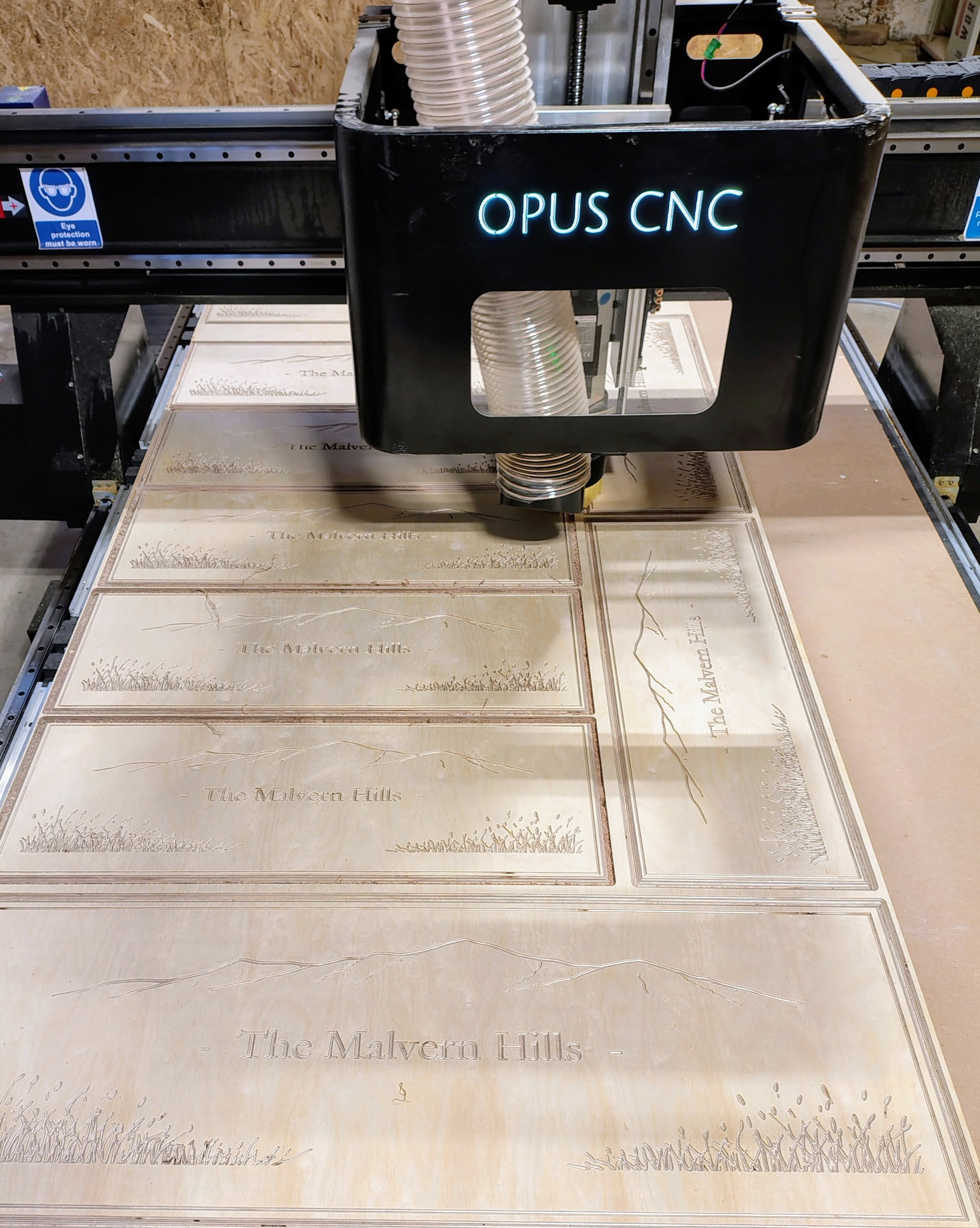 The image shows a CNC machine cutting tiles with the text "The Malvern Hills" visible and a black illuminated sign with the words "OPUS CNC". The tiles have a design resembling a map or landscape with detailed text and lines.